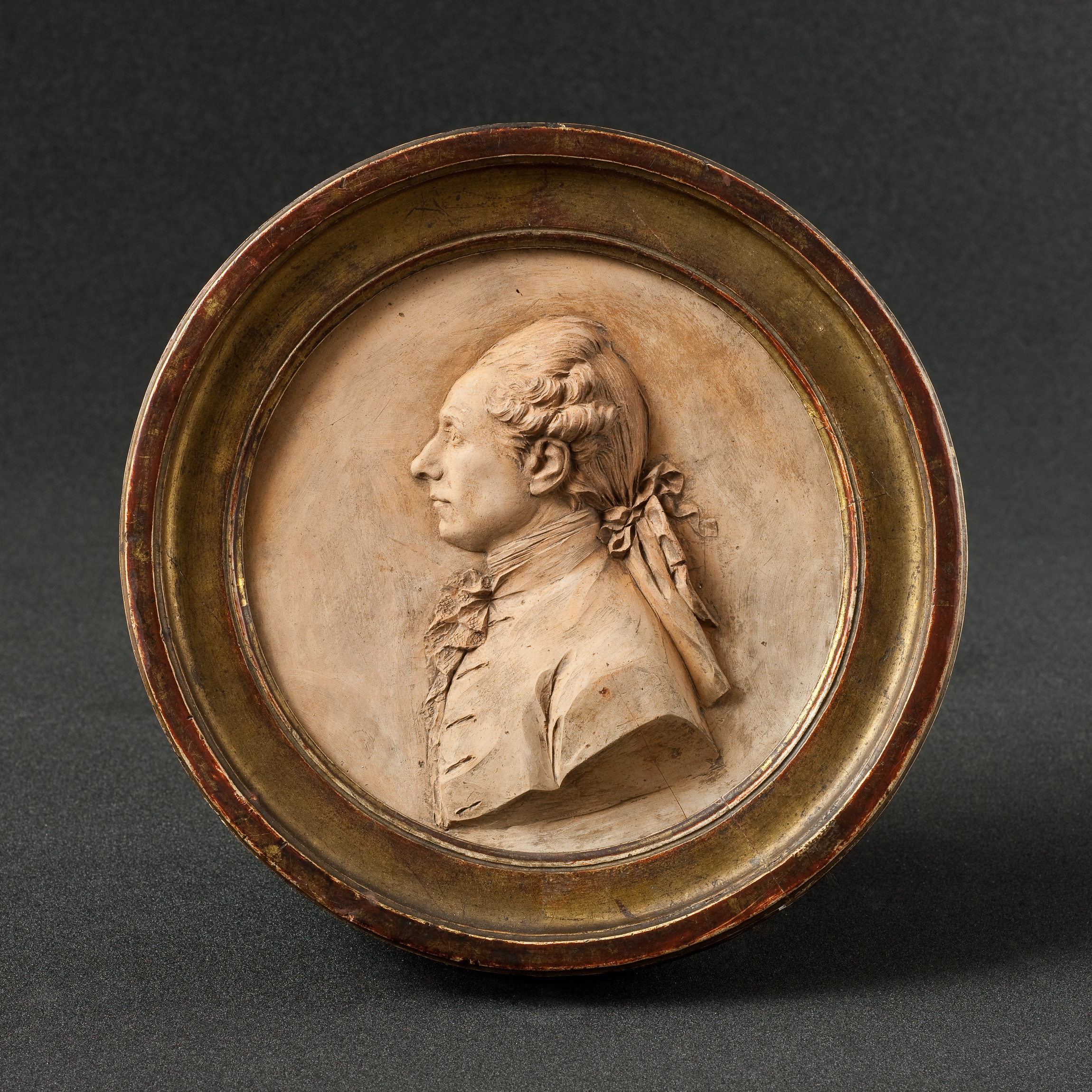 L'image montre un médaillon en relief représentant un profil. Le personnage est vêtu d'un habit d'époque, avec des détails soignés sur les vêtements. Ses cheveux sont coiffés de manière élégante et il porte un nœud à l'arrière de la tête. Le fond du médaillon est lisse, créant un contraste avec la texture du relief. L'ensemble est entouré d'une bordure circulaire, suggérant une finition soignée et un aspect décoratif. 