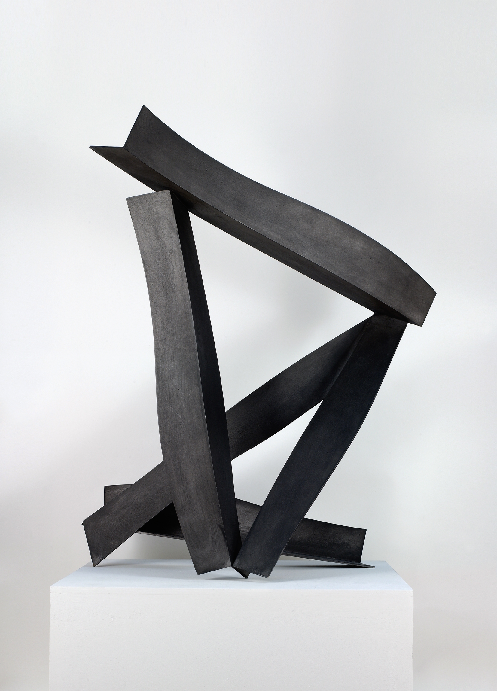 L'image montre une sculpture moderne en métal, composée de plusieurs éléments rejoignant des formes géométriques. Les parties de la sculpture sont disposées de manière dynamique, avec des angles et des courbes qui créent une impression de mouvement. La finition est lisse et d'un noir profond, ce qui apporte une certaine élégance et une esthétique contemporaine à l'œuvre. Elle est placée sur un socle blanc, ce qui met en avant les contrastes entre la sculpture et son support.