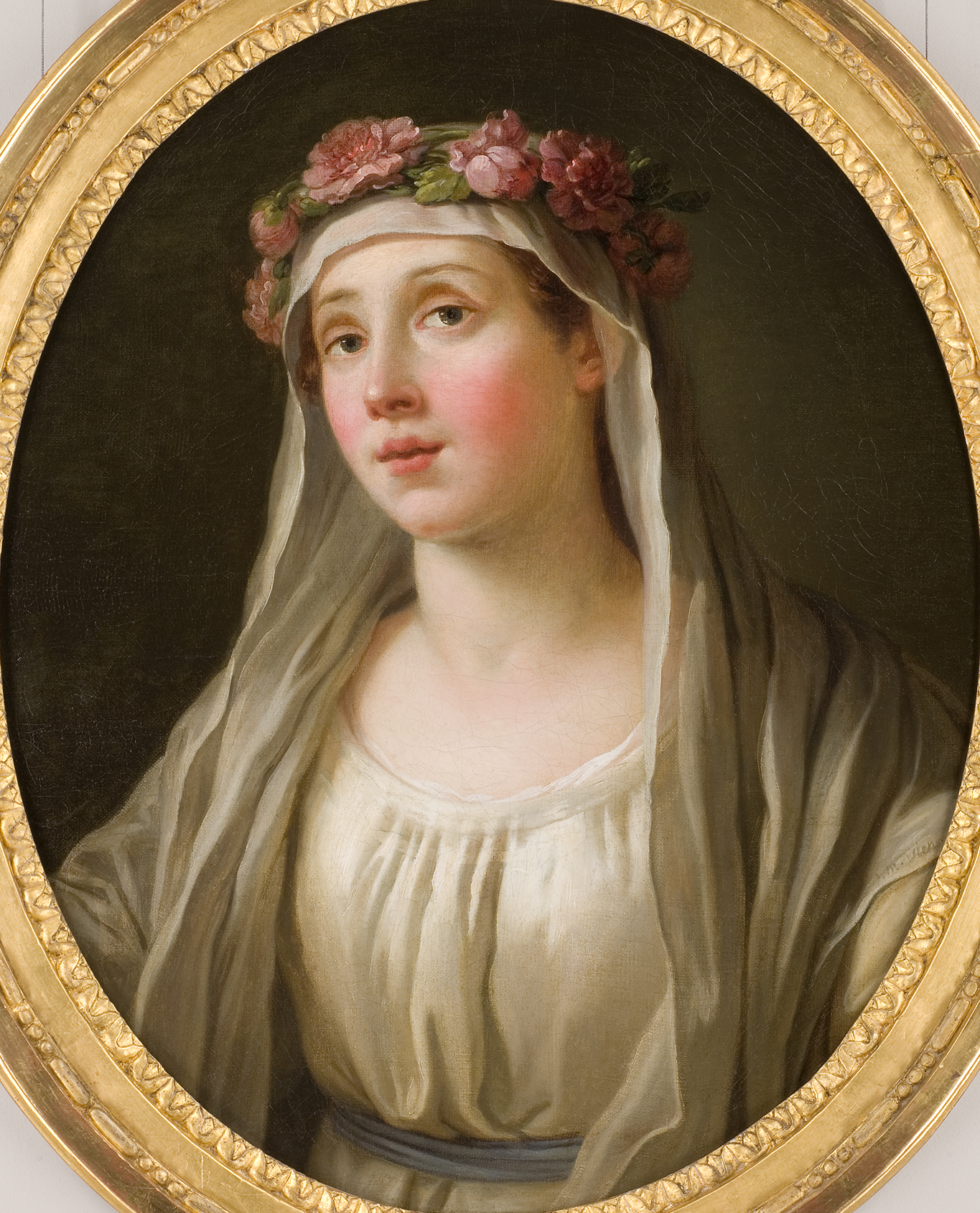L'image représente un portrait d'une jeune femme. Elle porte une couronne de fleurs sur la tête et a un regard doux et pensif. Son visage est légèrement rosé, et elle revêt une tenue simple, avec un décolleté discret. Un voile léger encadre son visage, ajoutant une touche de douceur à son apparence. Le fond est sombre, ce qui met en valeur les caractéristiques de son visage et les détails de sa tenue. Le cadre est orné, ajoutant une note d'élégance à l'ensemble.