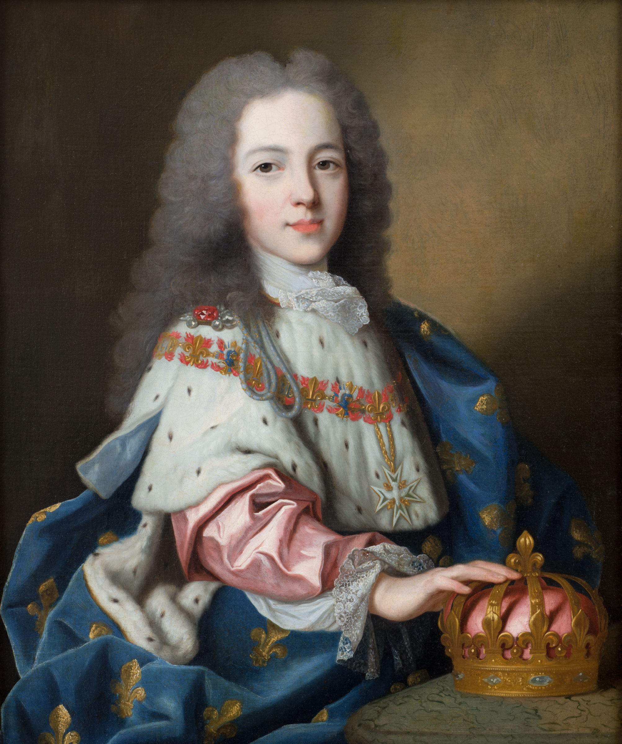 L'image représente un jeune garçon portant une riche tenue royale, ornée de détails luxueux. Il est assis, avec une expression douce sur son visage. Son costume est composé d'une cape bleu royal, avec des motifs fleuris dorés, et il porte une chemise blanche sous la cape. On peut voir une couronne en or et en velours rouge posée sur la table devant lui. Le décor est élégant et reflète une ambiance de grandeur royale, mettant en valeur le statut et l'importance de la personne représentée.