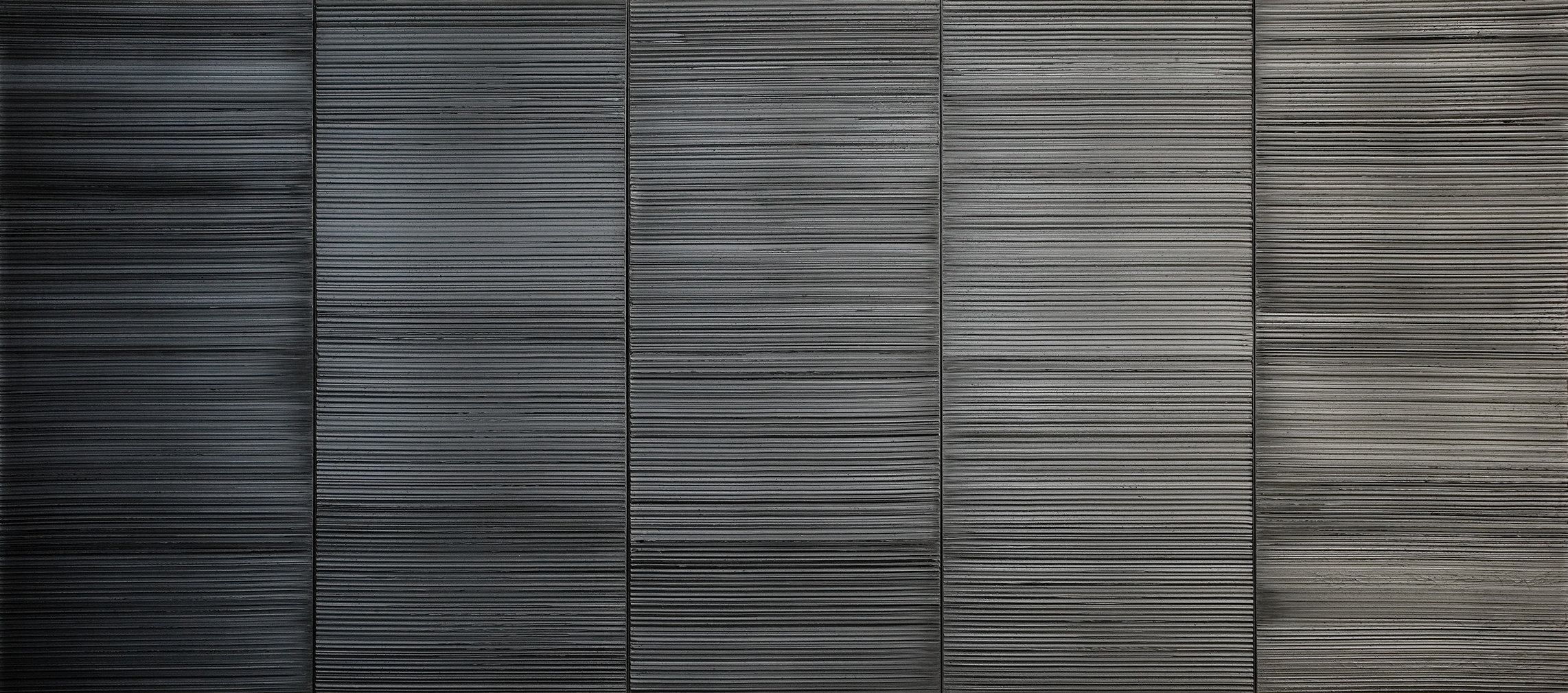 L'image montre une surface composée de plusieurs panneaux disposés verticalement. Chaque panneau présente une texture rayée, avec des lignes horizontales uniformes qui créent un effet de profondeur. Les nuances de gris varient légèrement d'un panneau à l'autre, allant du noir au gris clair, donnant une impression de modernité et de sophistication. L'ensemble de l'image dégage une atmosphère minimaliste et contemporaine.