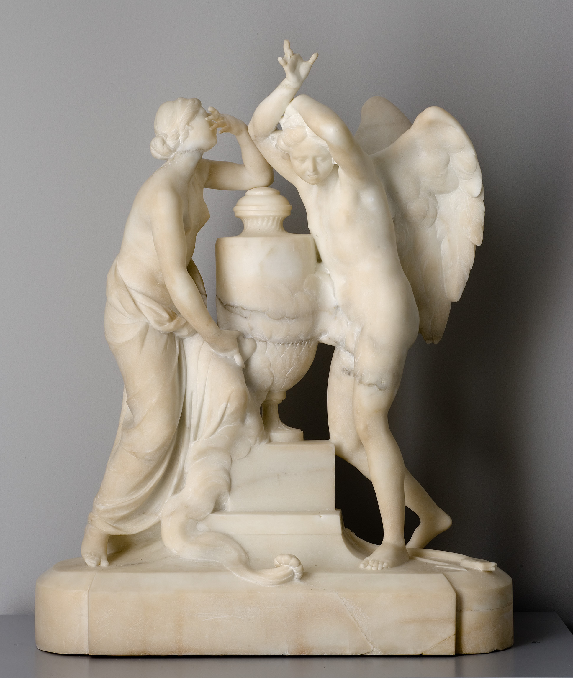 L'image représente une sculpture en marbre. On y voit deux figures humaines, une femme et un homme ailé, qui semblent interagir autour d'un urne. La femme, drapée dans un vêtement, se penche légèrement en avant, tandis que l'homme, qui a des ailes, adopte une posture dynamique, levant les bras. La sculpture est détaillée, mettant en valeur les expressions et les drapés des vêtements, créant une ambiance artistique et romantique. Les formes sont bien travaillées, ce qui confère une impression de mouvement et de vie à l'œuvre.