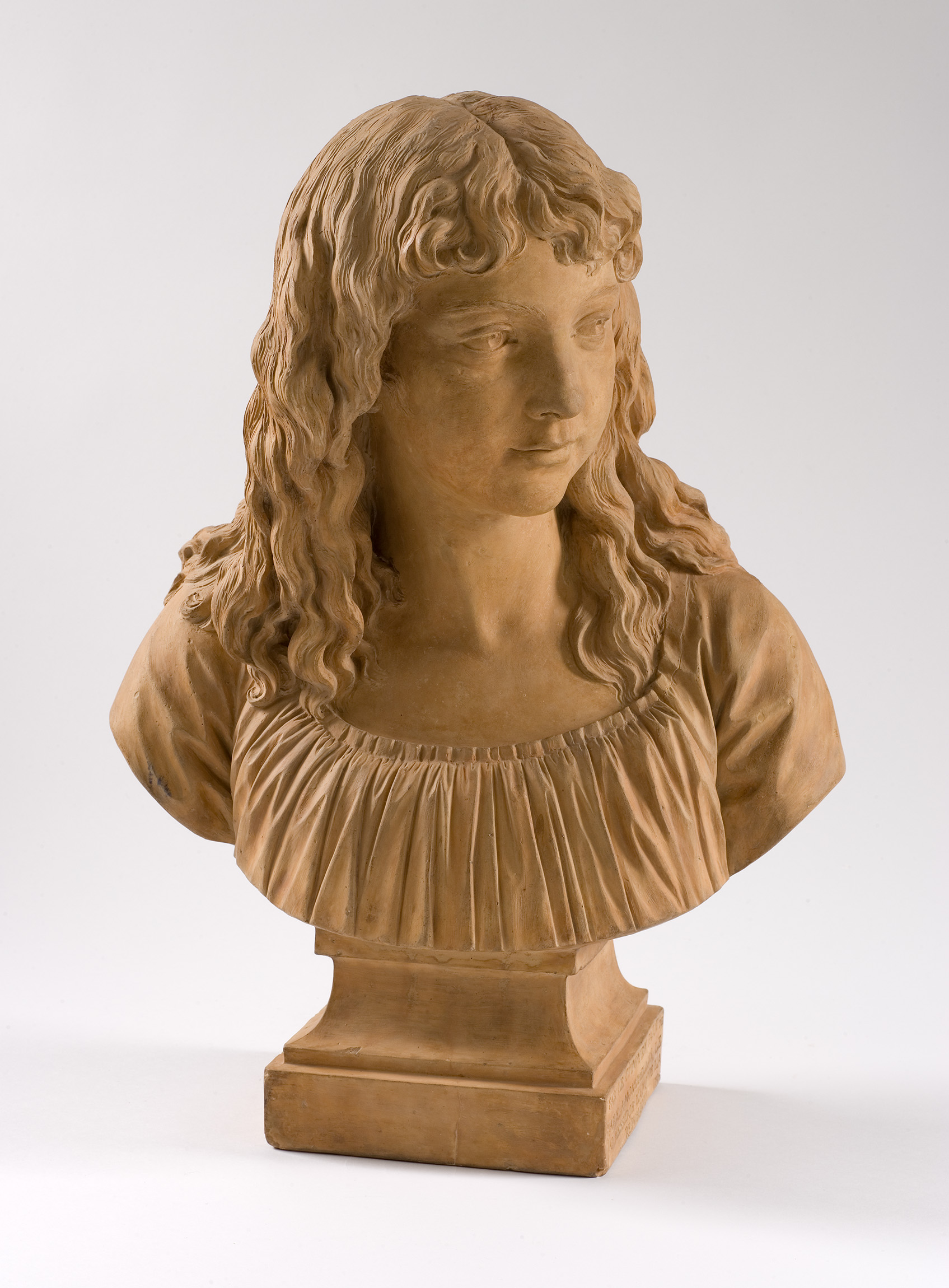 Cette image montre un buste en terre cuite représentant une jeune fille. La sculpture met en valeur les détails des cheveux longs et bouclés, ainsi que le visage doux et expressif de la fillette. Elle porte une blouse avec un décolleté plissé, et le buste repose sur un socle carré. La lumière éclaire délicatement les traits de la sculpture, accentuant les textures et les ombres. L'ensemble dégage une impression de sérénité et de grâce.