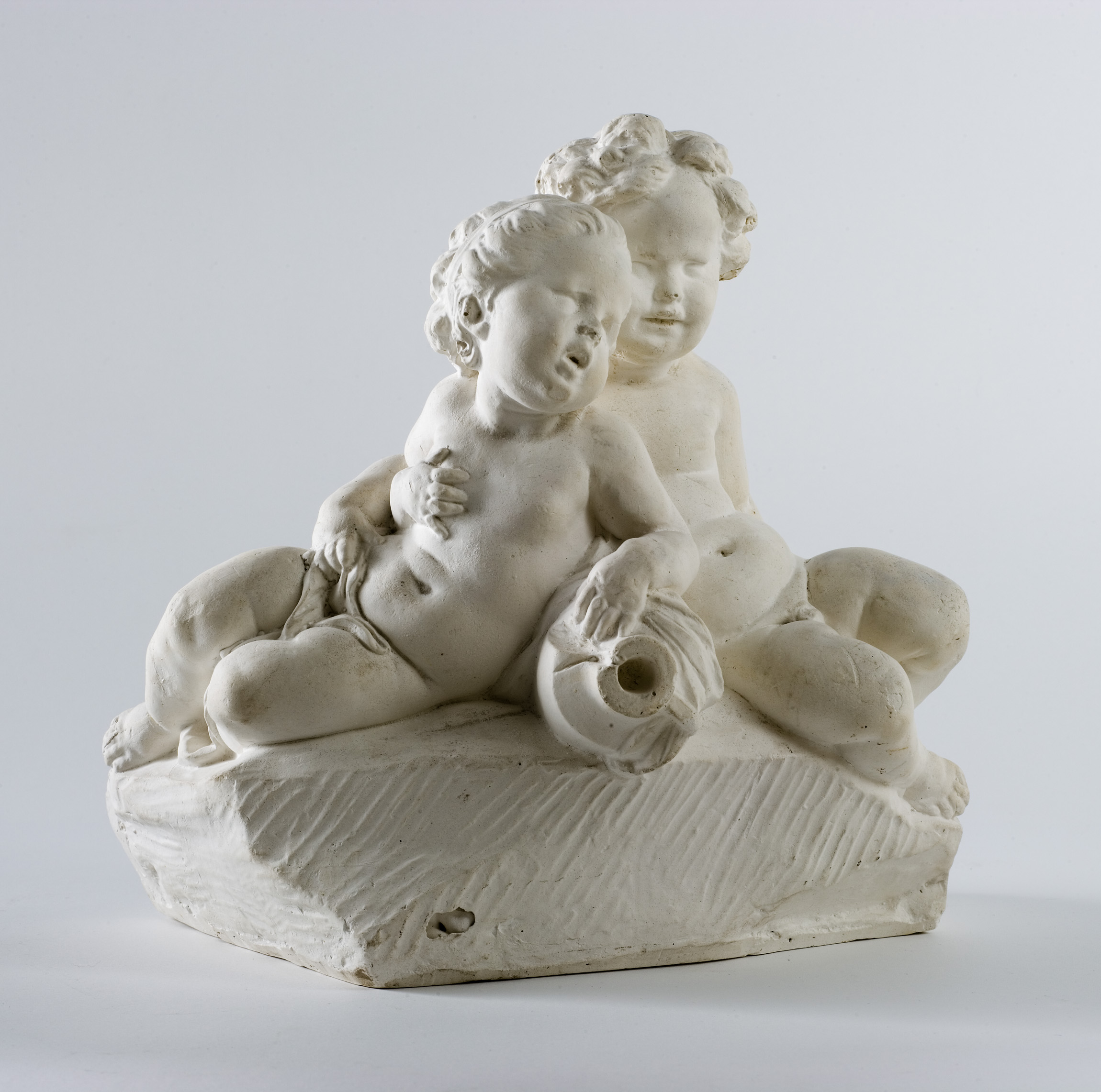 L'image montre une sculpture en plâtre représentant deux enfants nus assis côte à côte. Ils ont des cheveux bouclés et des expressions faciales douces. L'un des enfants tient un petit objet, tandis que l'autre semble penché vers lui. La sculpture est détaillée, montrant des traits délicats et une certaine rondeur dans les formes, le tout reposant sur une base irrégulière. Le fond est bien neutre, mettant en valeur la blancheur et la texture de l'œuvre.
