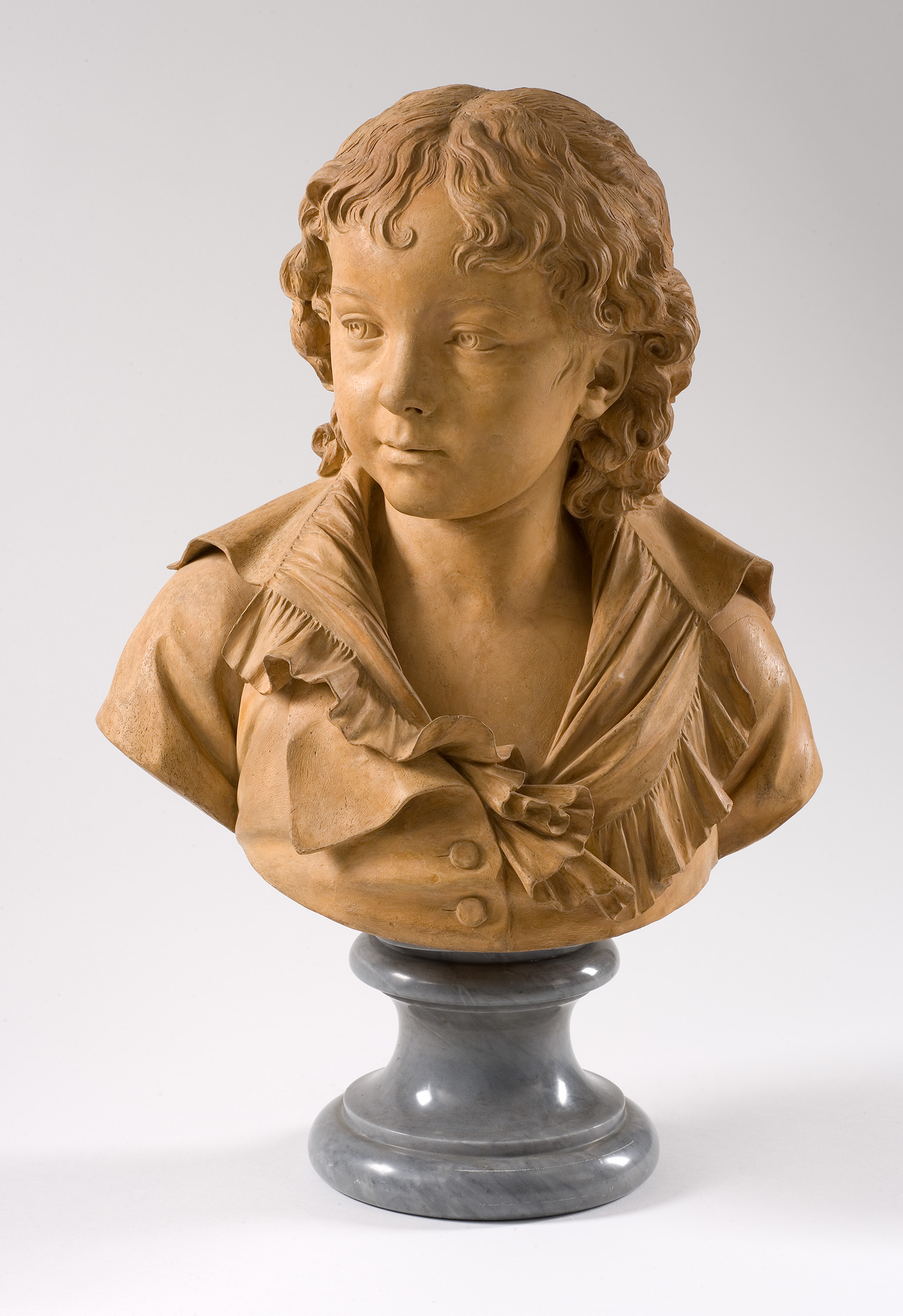 L'image représente un buste en terre cuite d'un jeune garçon. Il a les cheveux bouclés et porte un vêtement qui semble être une veste avec un col. Le détail des plis de la veste et des traits du visage est particulièrement bien travaillé. Le buste repose sur un socle en marbre, ajoutant à l'élégance de la sculpture. Le regard du garçon est attentif et innocent, ce qui donne une expression douce et captivante à l'œuvre.