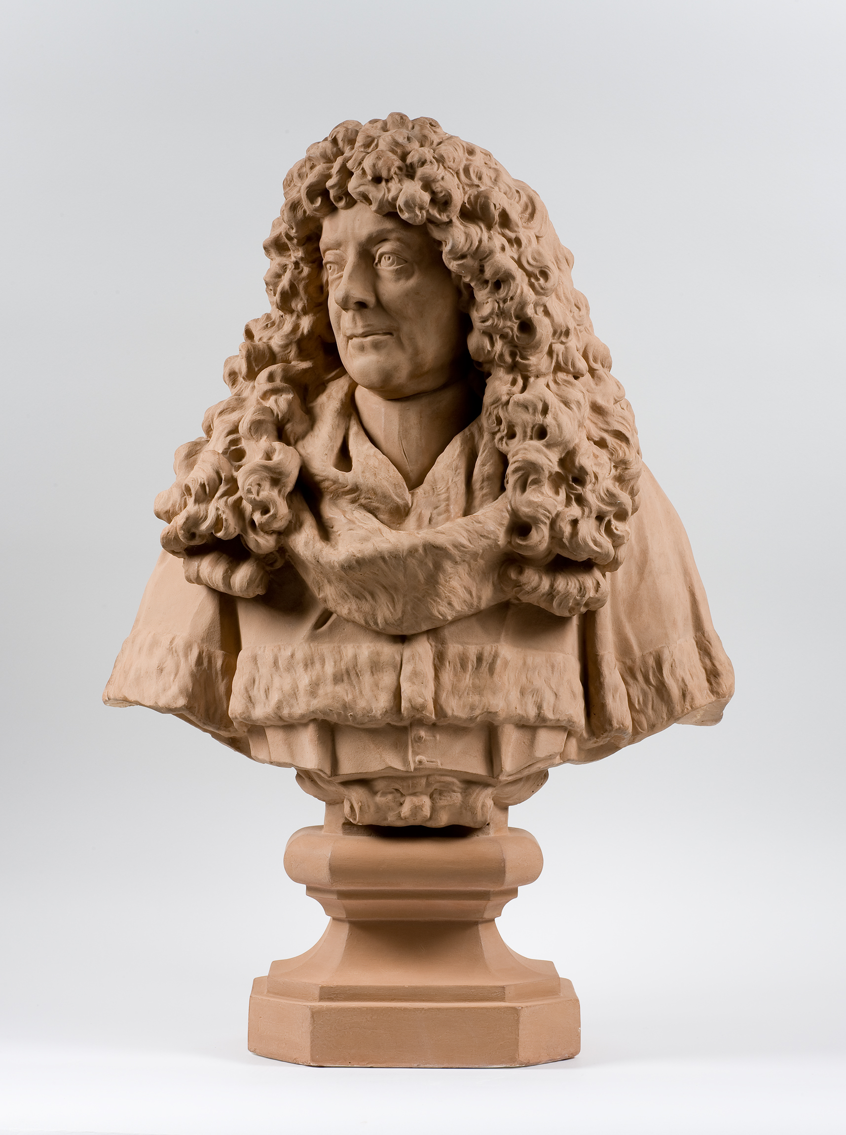 L'image présente une sculpture en terre cuite représentant un buste d'une personne avec de longs cheveux bouclés. La sculpture est détaillée, montrant des expressions faciales et des vêtements élaborés, tout en mettant en évidence le mouvement des cheveux. Le buste est posé sur un socle, ce qui lui confère une certaine élévation et importance. L'œuvre semble être à la fois réaliste et artistique.