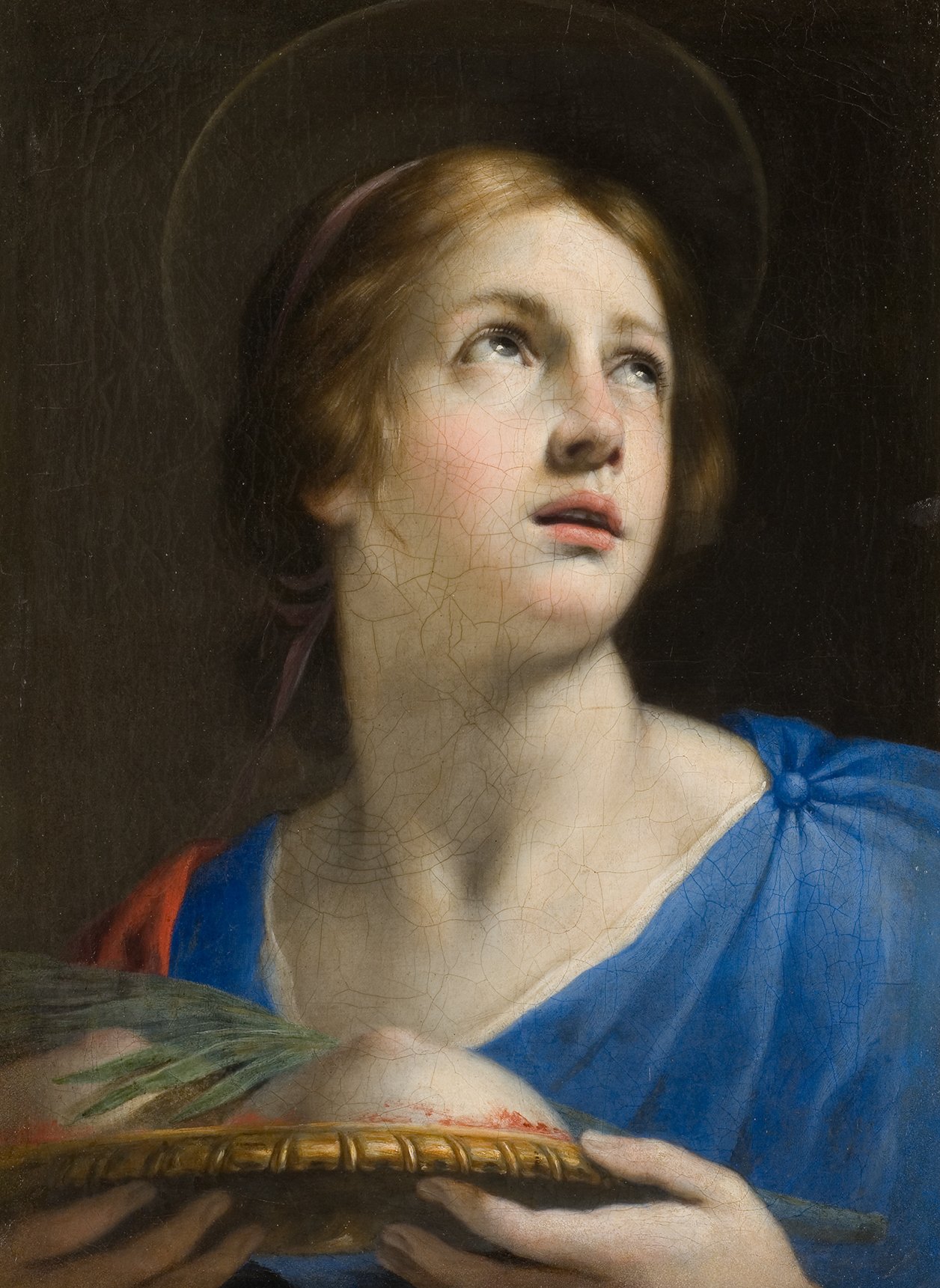 L'image représente une jeune femme au visage doux, semblant contempler le ciel ou se perdre dans ses pensées. Elle porte une robe bleue avec un drapé rouge et a des cheveux blonds encadrant son visage. Sa posture indique qu'elle tient un plat ou un plateau, souvent associé à des thèmes religieux ou symboliques. Un halo autour de sa tête suggère une aura de sainteté. L'arrière-plan sombre met en valeur la luminosité de son visage et sa tenue.