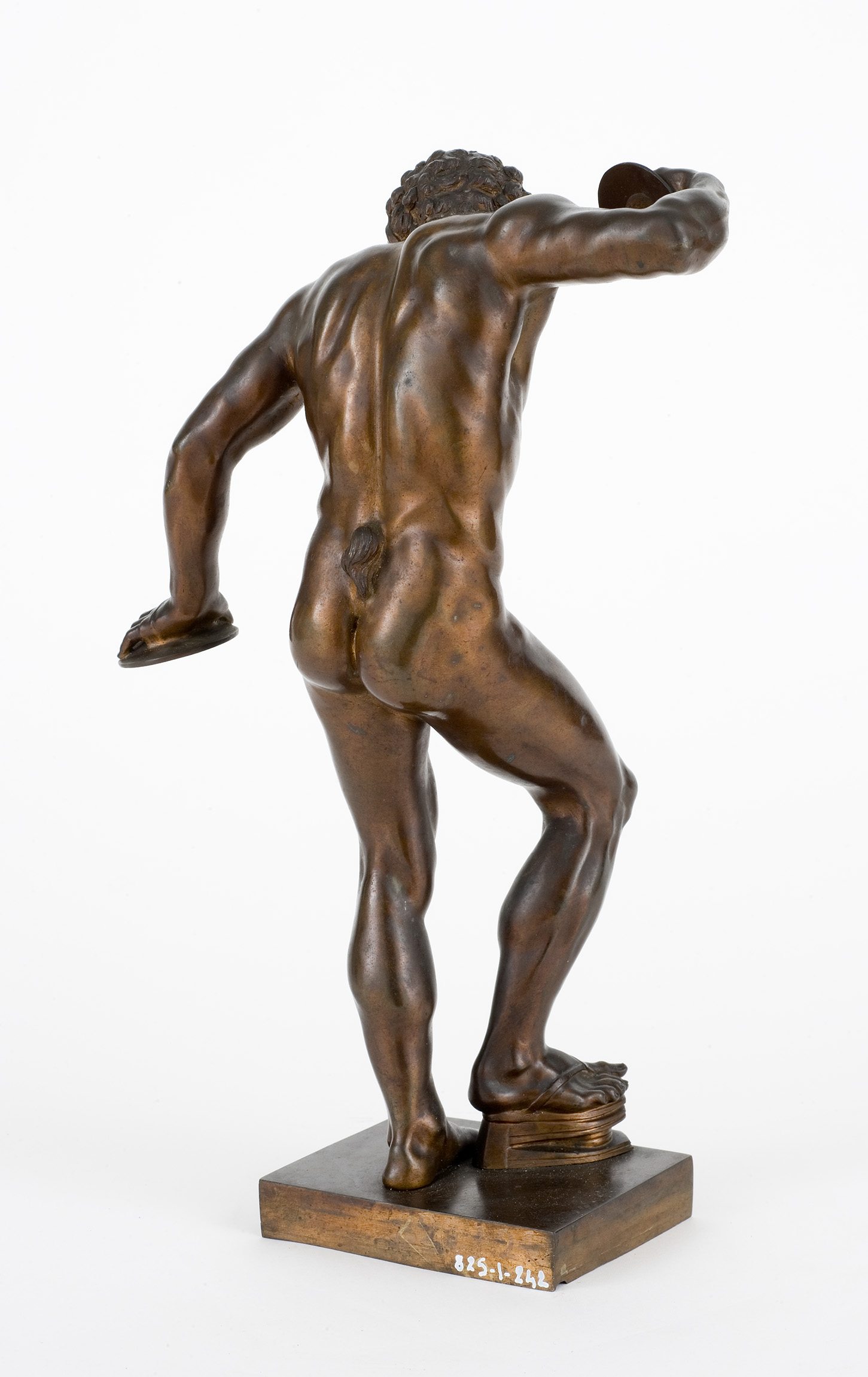 L'image représente une sculpture en bronze d'un athlète à la musculature développée, vu de dos. Il est en position dynamique, comme s'il était en train de lancer un disque. La sculpture met en valeur les détails anatomiques, notamment les muscles des bras et des jambes. La base de la statue est carrée et semble bien travaillée. L'ensemble dégage une impression de force et de mouvement.