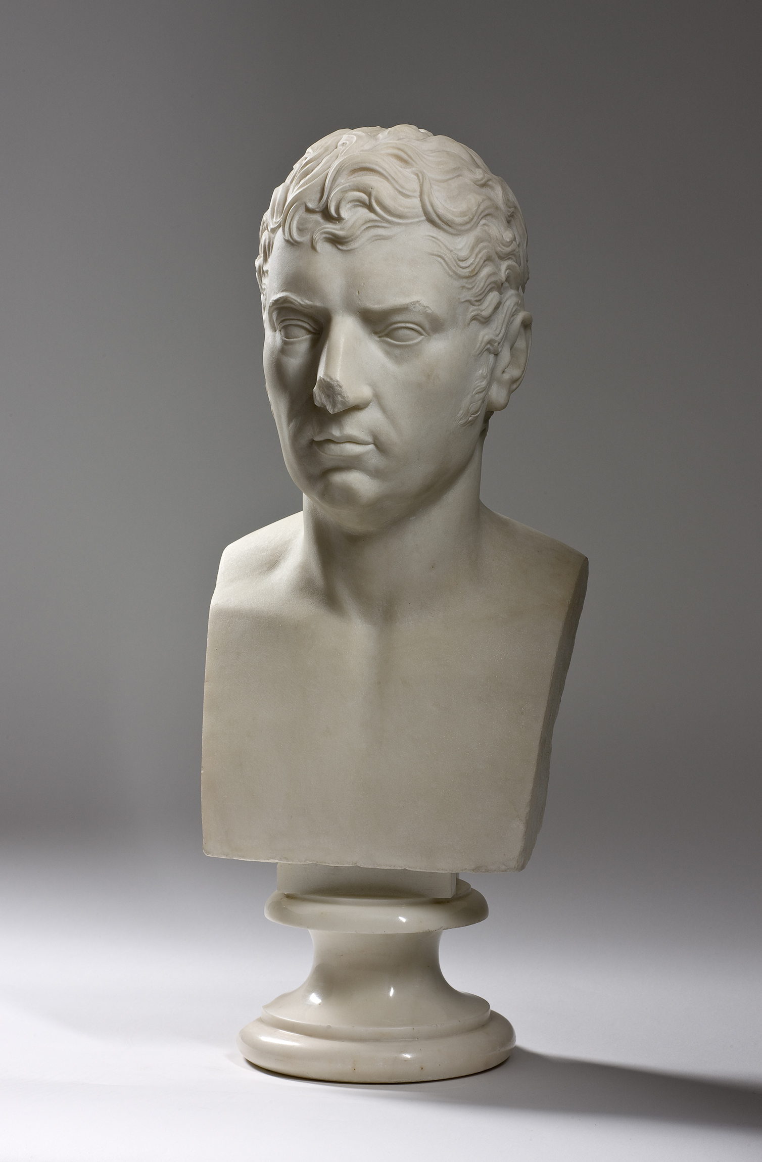 L'image montre un buste en marbre représentant un jeune homme. Le buste est sculpté avec des détails soignés, mettant en avant des traits du visage tels que des lèvres fines et des cheveux ondulés. Il est placé sur un socle circulaire, ce qui lui donne une certaine élégance. Le fond est neutre, ce qui permet de mettre en valeur la sculpture. Le visage exprime une certaine intensité, avec un regard sérieux et pensif.