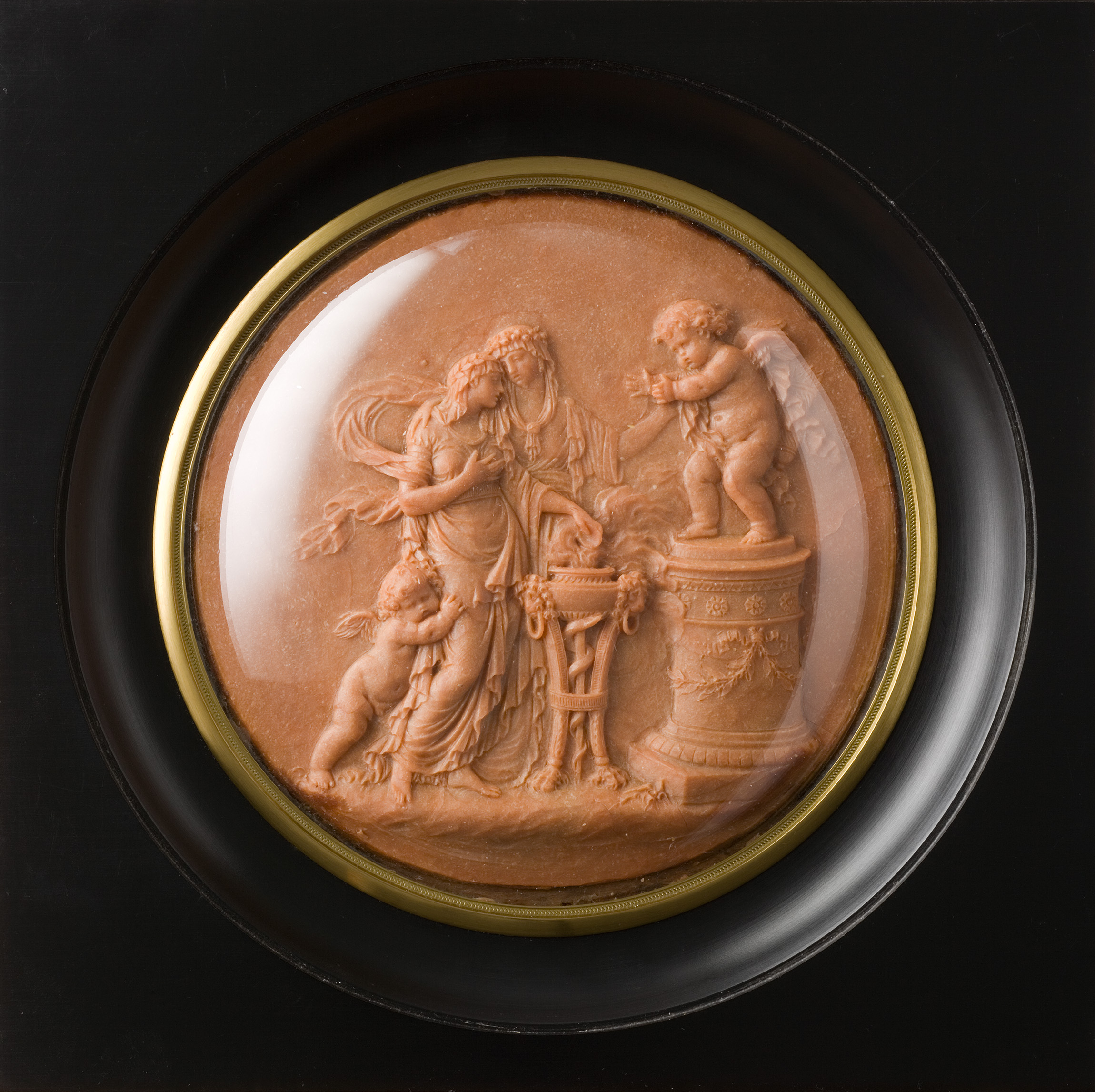 Cette image présente une œuvre d'art en relief, probablement un médaillon ou un bas-relief, qui représente une scène mythologique ou allusive. Au centre, on peut voir une figure féminine qui semble incarner une déesse ou une allégorie, entourée par des putti (petits anges) qui interagissent avec elle. L'un des putti est debout sur un socle, tandis que l'autre, plus petit, est à ses côtés. La composition évoque une atmosphère douce et élégante, avec des détails délicats qui illustrent les sentiments de tendresse et de douceur. Le fond est circulaire et encadré, ce qui met en valeur la scène au centre.