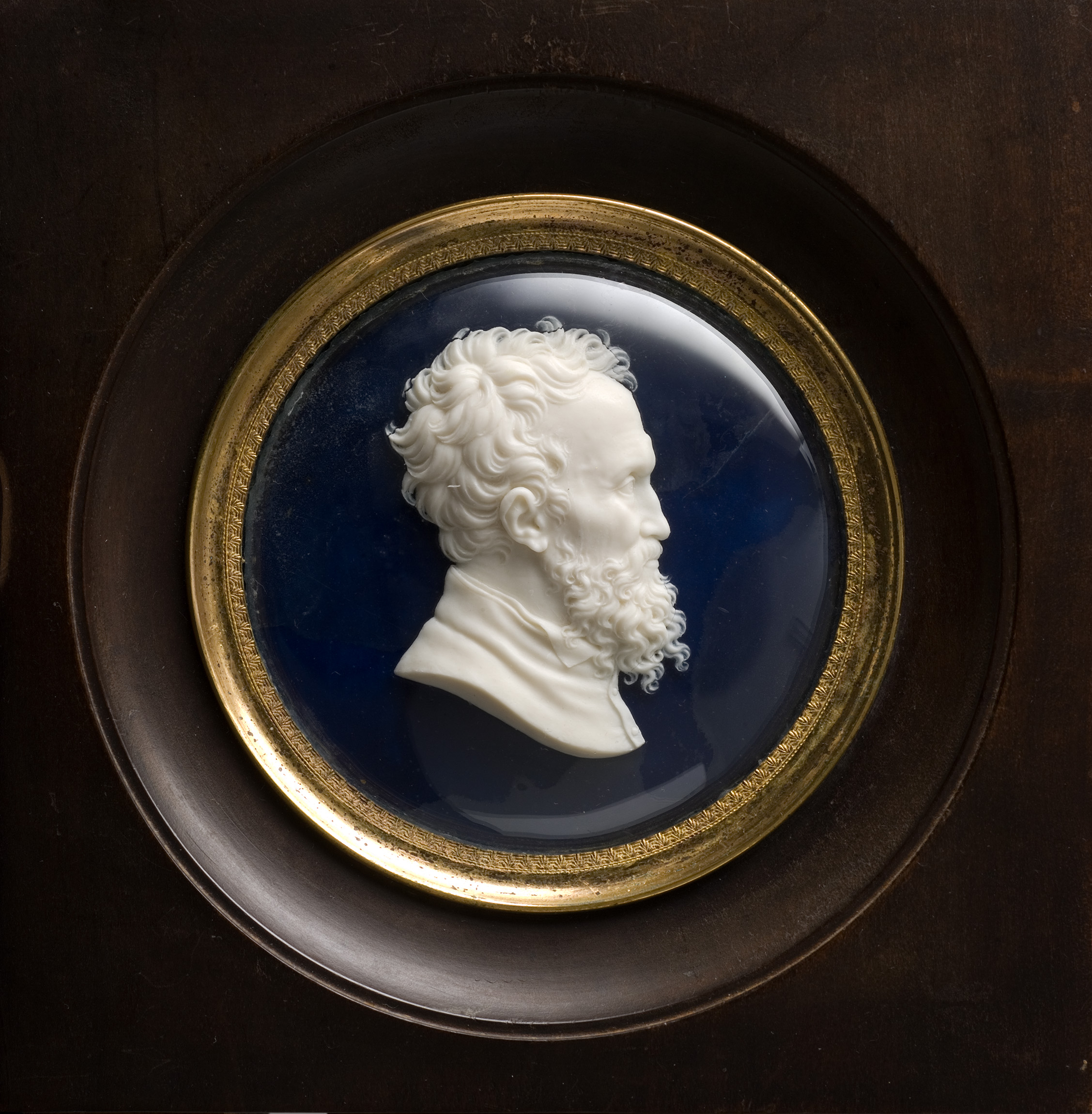 L'image représente un médaillon en relief, mettant en avant un portrait en profil d'un homme. Ce dernier a une chevelure et une barbe ondulées, ce qui lui confère une apparence distinguée. Le fond du médaillon est de couleur bleu profond, et le tout est encadré par un cercle doré. Le médaillon est intégré dans un cadre sombre qui accentue le contraste entre les différentes couleurs et matériaux.