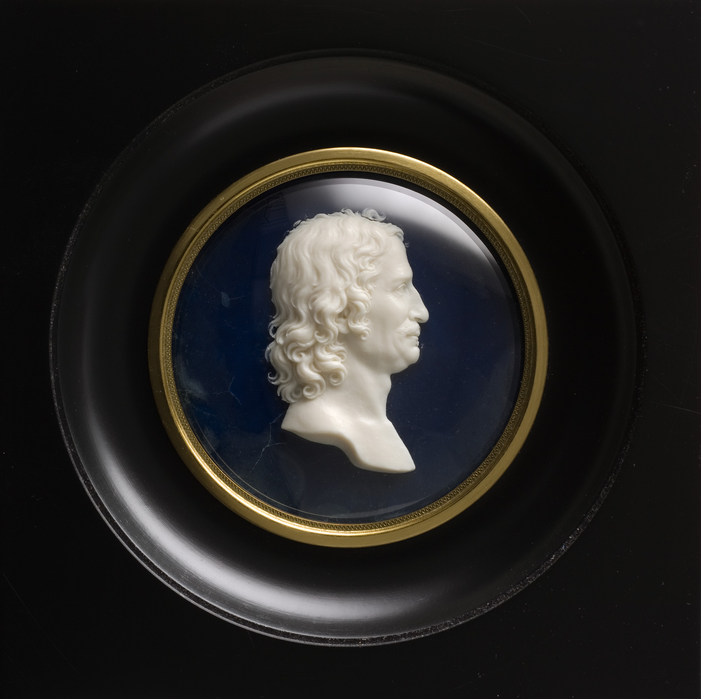 L'image présente un médaillon, dans un cadre circulaire noir, contenant une sculpture en relief d'un profil masculin. La sculpture, probablement en porcelaine ou en ivoire, montre des détails fins, tels que les cheveux bouclés et les traits du visage. Le fond du médaillon est bleu, et il est entouré d'un liseré doré, ajoutant à l'élégance de la pièce. Cette œuvre évoque le style néoclassique, caractérisé par un souci du détail et une esthétique classique.