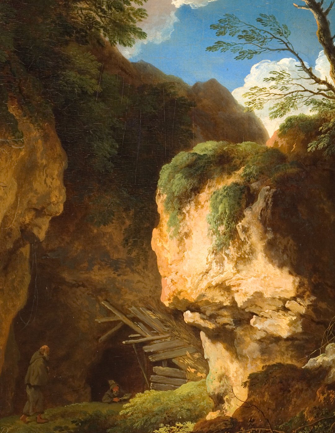 L'image montre un paysage naturel avec une grotte partiellement cachée derrière des rochers. La lumière du jour éclaire la scène, mettant en valeur le vert des plantes et la texture des pierres. On aperçoit également deux personnages : l'un semble se tenir près de l'entrée de la grotte, tandis que l'autre est plus éloigné. Le ciel est bleu avec quelques nuages, ce qui ajoute à l'ambiance sereine de l'environnement. L'ensemble de la composition évoque une atmosphère paisible et mystérieuse.