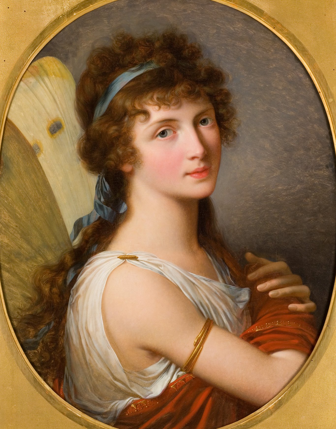 L'image représente un portrait d'une jeune femme, affichant des traits délicats et une expression douce. Elle porte une robe blanche ornée de draperies rouges et un bandeau bleu dans ses cheveux bruns légèrement ondulés. Son regard paisible est dirigé vers le spectateur, et elle repose ses bras croisés sur sa poitrine. Le fond de l'image est flou et met en valeur le visage et les vêtements de la femme, créant une atmosphère élégante et artistique. Entouré d'une bordure dorée, le portrait dégage une impression de finesse et de raffinement.