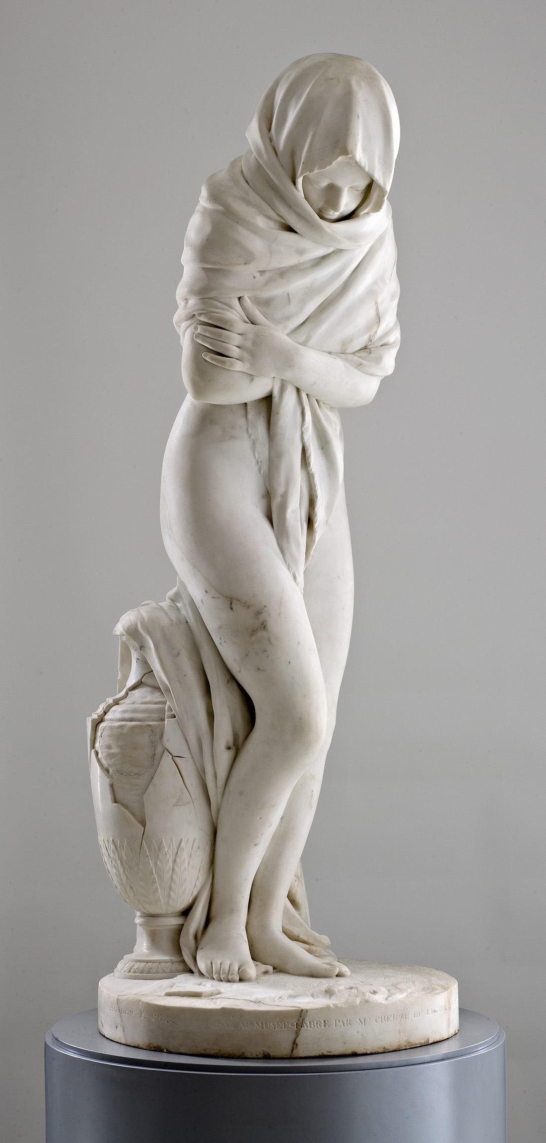 Cette image représente une sculpture en marbre d'une femme debout, légèrement courbée. Elle est partiellement drapée dans un voile qui recouvre ses épaules et ses bras, tout en laissant ses jambes nues visibles. La femme semble pensive et réservée, se tenant dans une posture protectrice. À ses pieds, on aperçoit un vase orné, ajoutant une dimension artistique à l'œuvre. La sculpture présente des détails délicats et raffinés, mettant en avant la beauté et la grâce du mouvement.