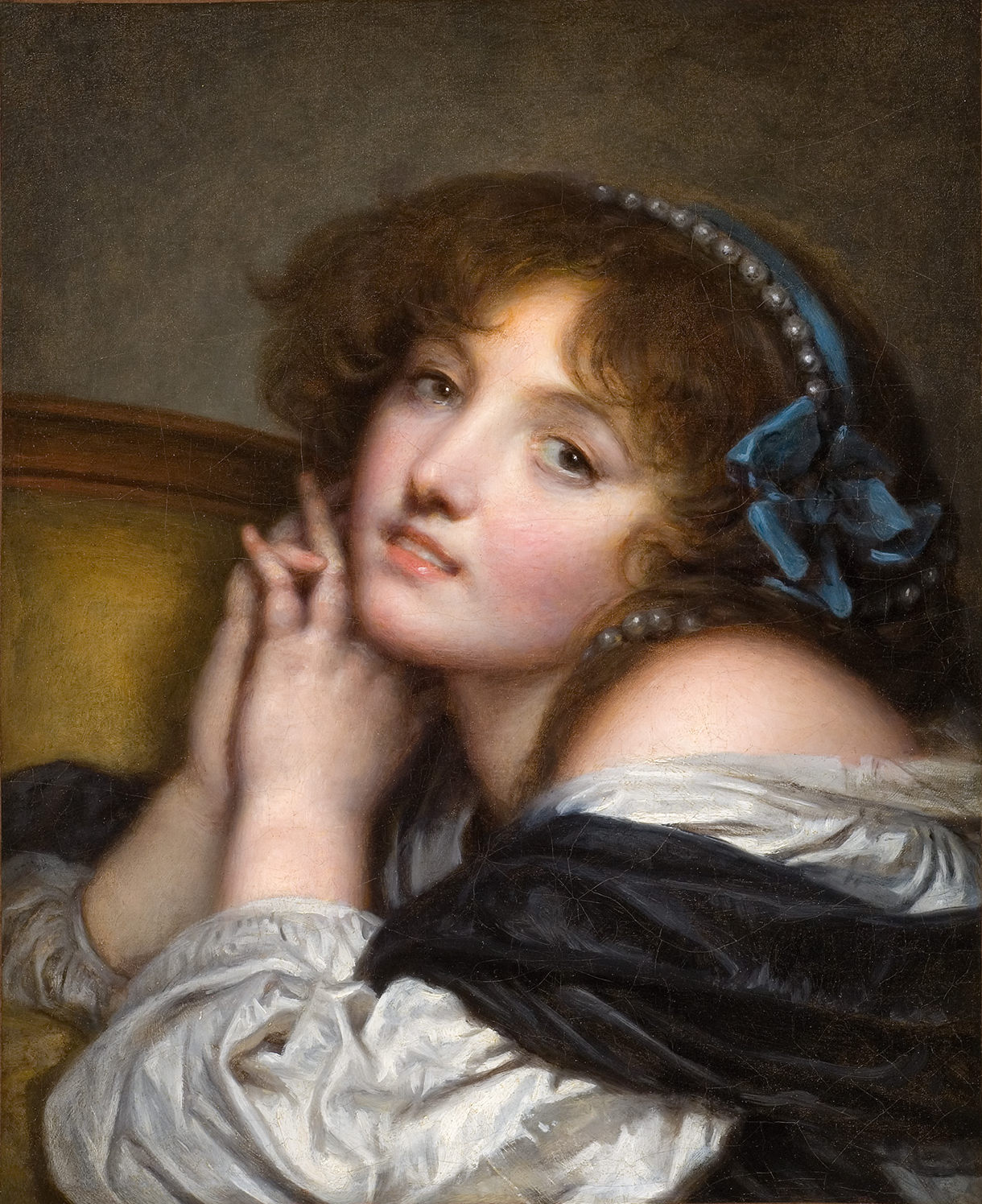 L'image représente un portrait d'une jeune femme avec des cheveux bouclés et un regard doux. Elle est vêtue d'une robe élégante, dont les manches sont en contraste avec une étole sombre. Sa main est délicatement posée sur son visage, tandis que l'autre main repose sur une surface. Un bandeau bleu et des perles ornent sa coiffure, ajoutant une touche de sophistication à son apparence. Le fond est sombre, ce qui met en valeur son visage et son expression.