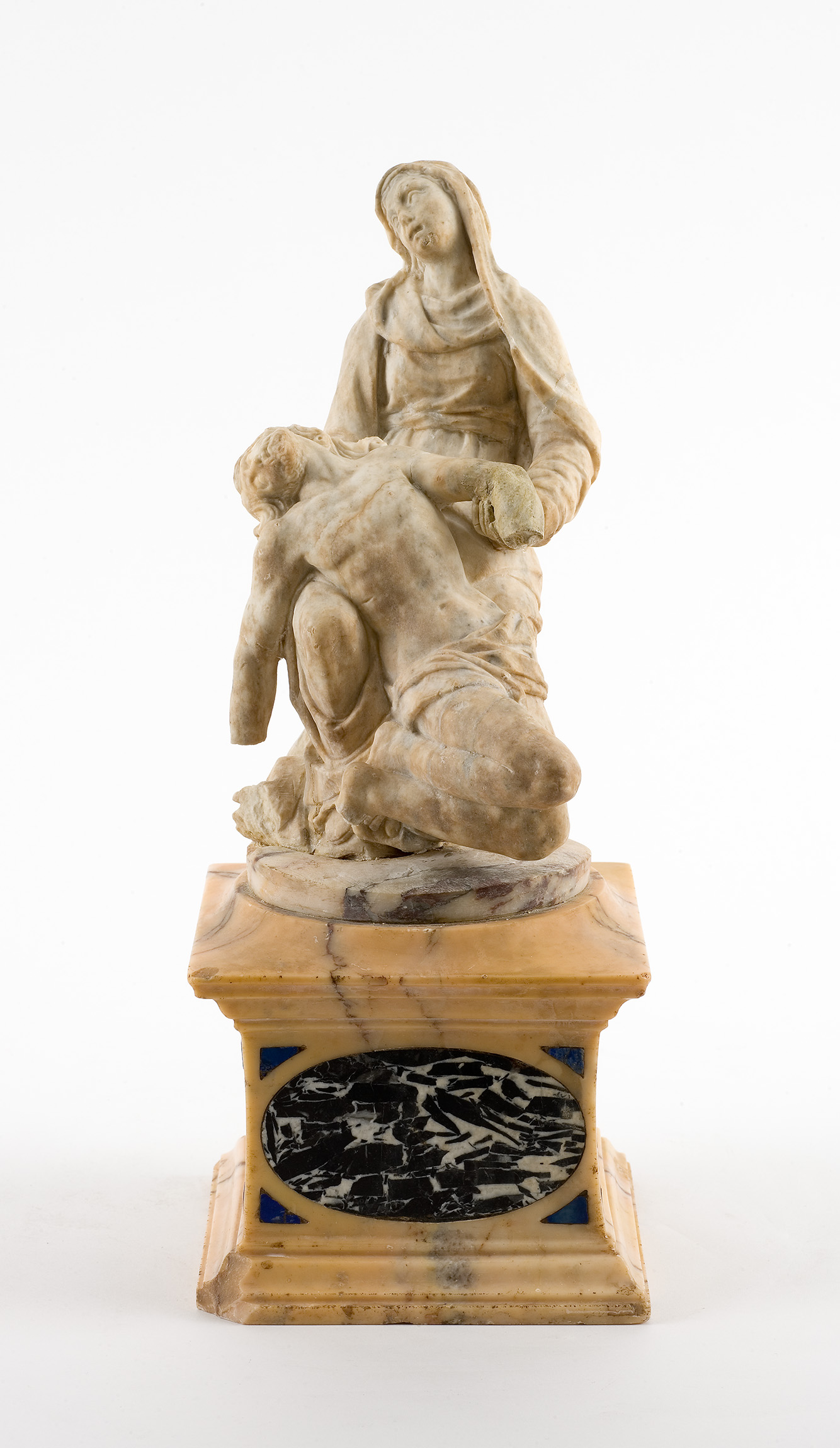 Cette image représente une sculpture en marbre, probablement une Pietà, où la Vierge Marie est assise, tenant le corps du Christ après la crucifixion. Marie a une expression de tristesse et de compassion alors qu'elle regarde le corps de son fils. Le socle est décoré de marbre aux nuances dorées et comprend des éléments en marbre noir et des motifs. L'ensemble évoque une profonde émotion et une grande maîtrise artistique.