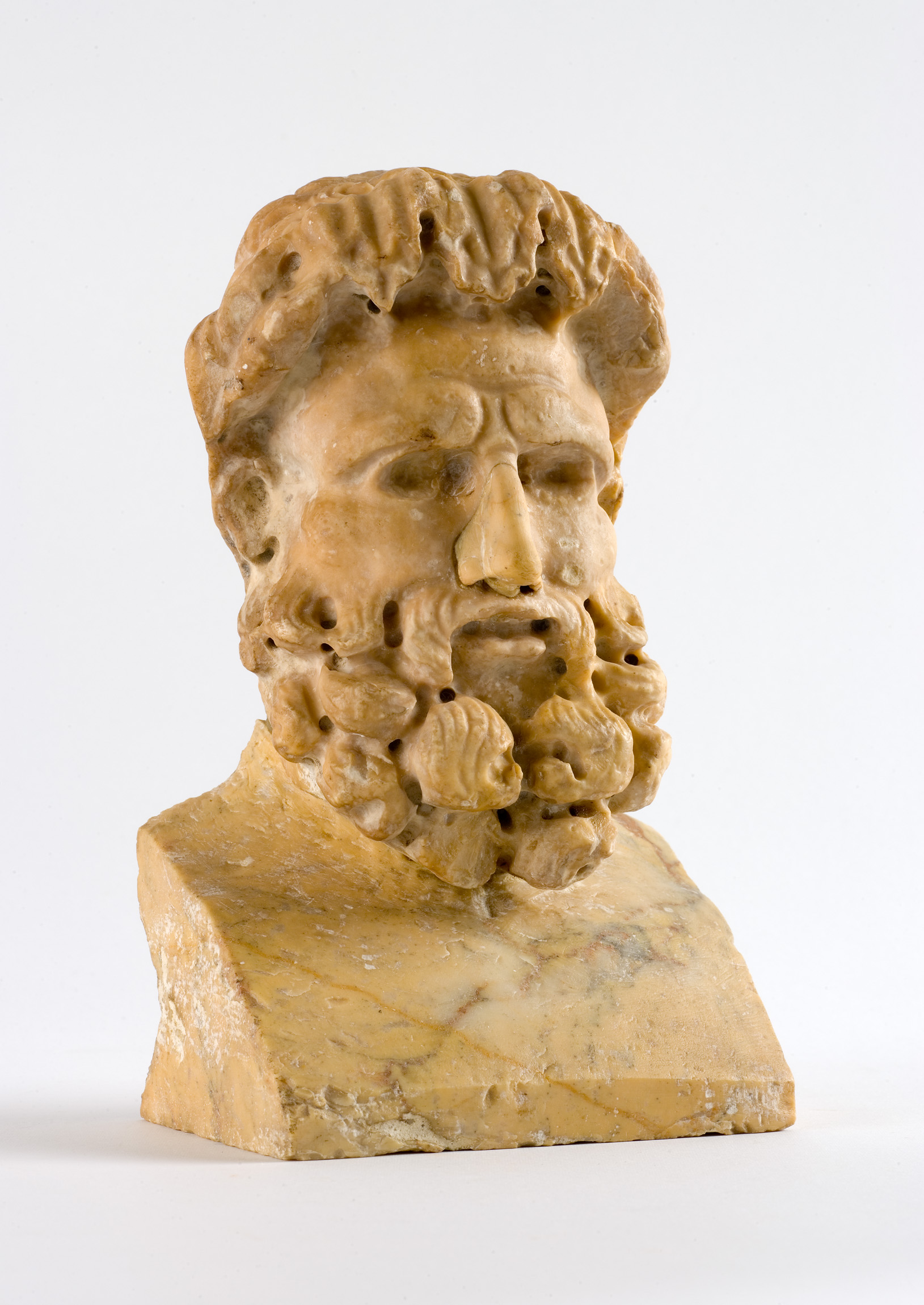 L'image montre une sculpture en marbre représentant un buste masculin. Le personnage a une barbe fournie et des cheveux ondulés. Sa expression est sérieuse, avec des traits du visage bien définis, notamment un nez prononcé et des lèvres closes. Le buste repose sur une base solide, mettant en valeur les détails sculptés. L'éclairage accentue les textures de la pierre, donnant une impression de profondeur et de réalisme à l'œuvre.