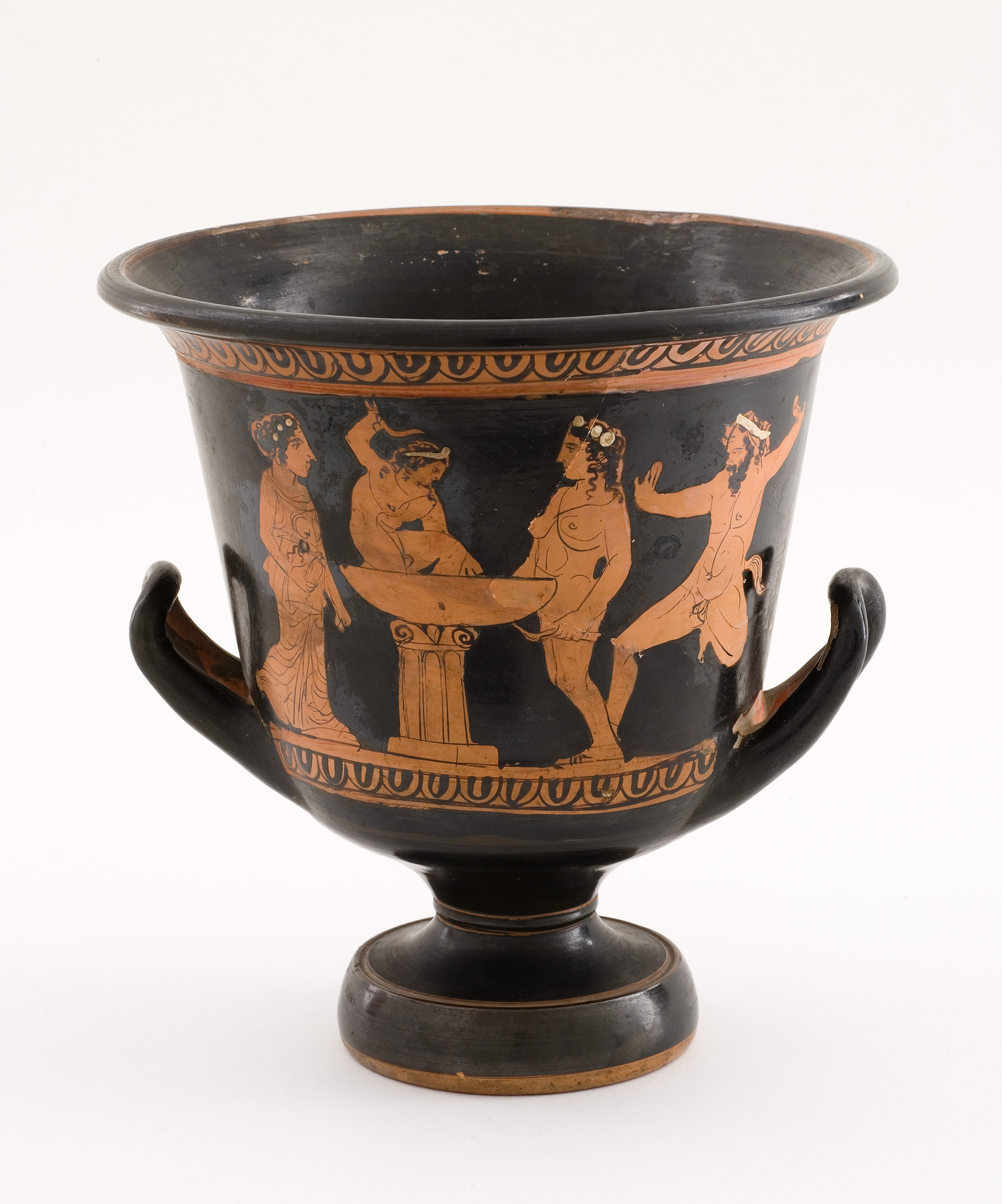 L'image montre un vase de style grec antique, spécifiquement une coupe appelée "kylix". Le vase est de couleur noire avec des dessins en reliefs orangés. On peut y voir des figures humaines, probablement en train de célébrer ou de danser, entourées de motifs floraux. Les personnages sont probablement des représentations de divinités ou de participants à des festivités, illustrant des scènes de joie et de convivialité. Les poignées et la base du vase sont soigneusement sculptées, ajoutant une touche d'élégance à l'ensemble.