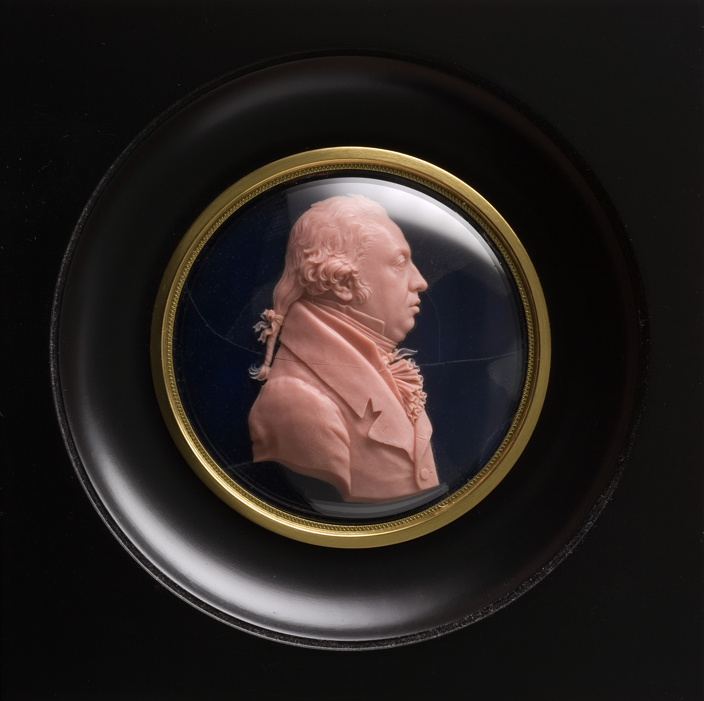 L'image représente un médaillon ovale en relief, mettant en avant un profil de personnage. Le modèle est sculpté dans une matière rose pâle, avec des détails fins, illustrant le visage d'un homme avec des cheveux coiffés en arrière et portant un vêtement élégant. Ce médaillon est encadré d'un bord doré et reposé sur un fond noir, ce qui met en valeur le contraste entre le rose du relief et le noir du fond. L'ensemble dégage une impression de sophistication et d'artisanat raffiné.