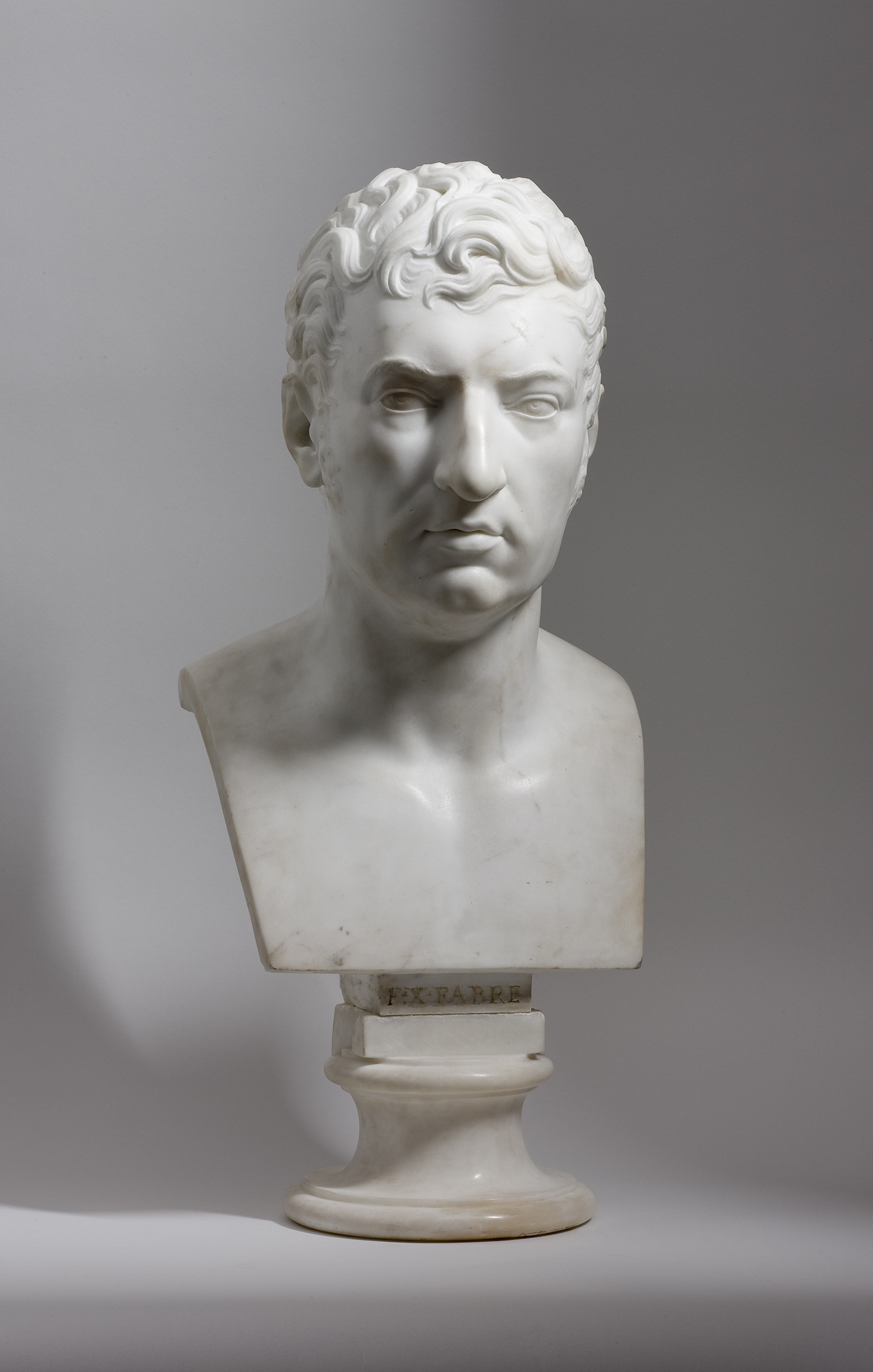 L'image montre un buste en marbre blanc représentant un homme avec des traits distincts. Il a des cheveux ondulés et une expression sérieuse. Le buste est sculpté de manière détaillée, mettant en avant les caractéristiques du visage, telles que le nez, les lèvres et les yeux. Il est monté sur un socle circulaire. L'arrière-plan est sobre, ce qui met en valeur le buste.