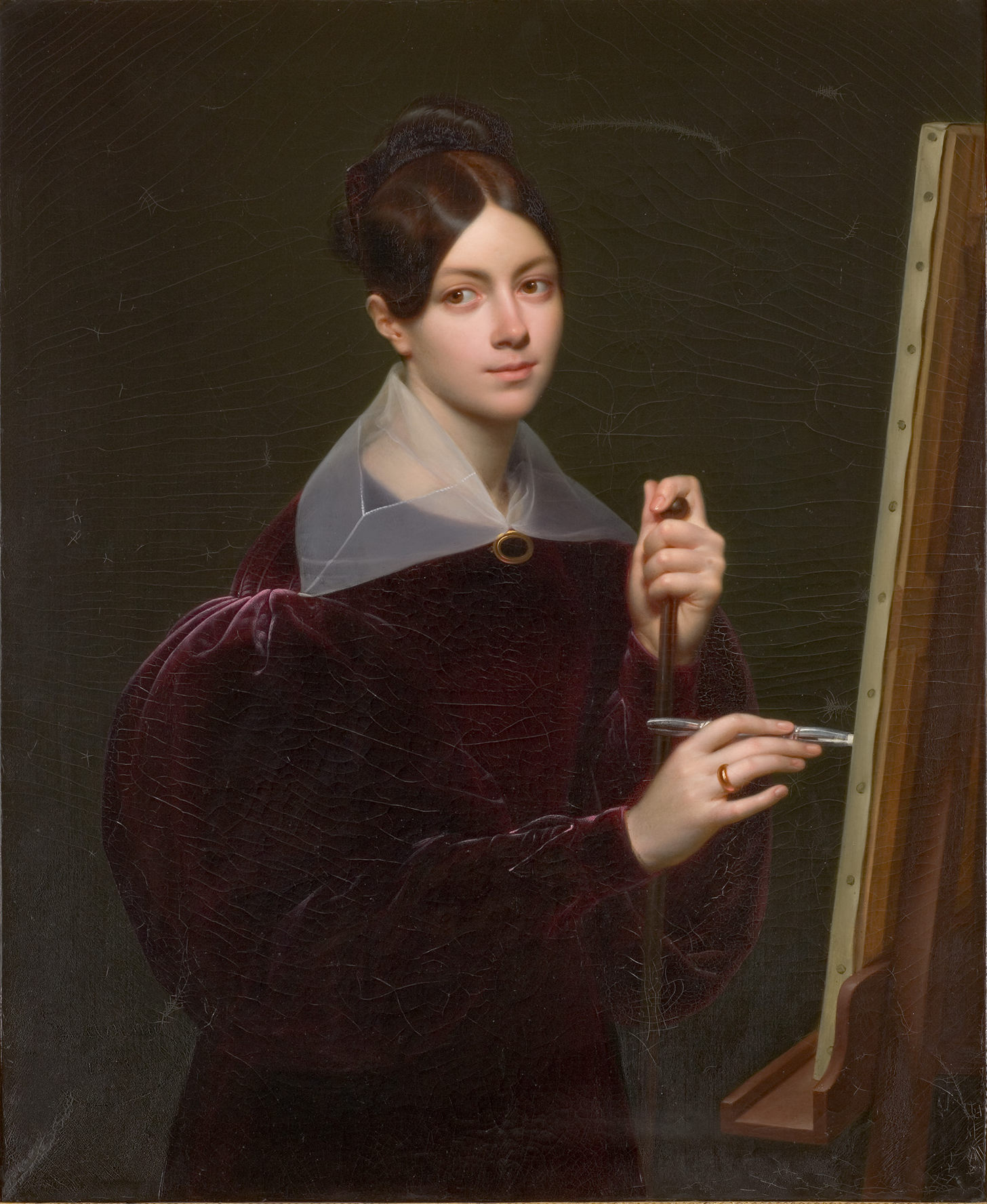 L'image représente une jeune femme de manière élégante, vêtue d'une robe en velours de couleur bordeaux. Elle tient un pinceau et se trouve devant un chevalet, semblant prête à peindre. Sa coiffure est soignée, avec un chignon, et elle porte un col en dentelle. L'expression sur son visage dénote à la fois concentration et douceur, permettant de saisir son engagement artistique. L'arrière-plan est sombre, ce qui met en valeur la luminosité de sa tenue et son visage.