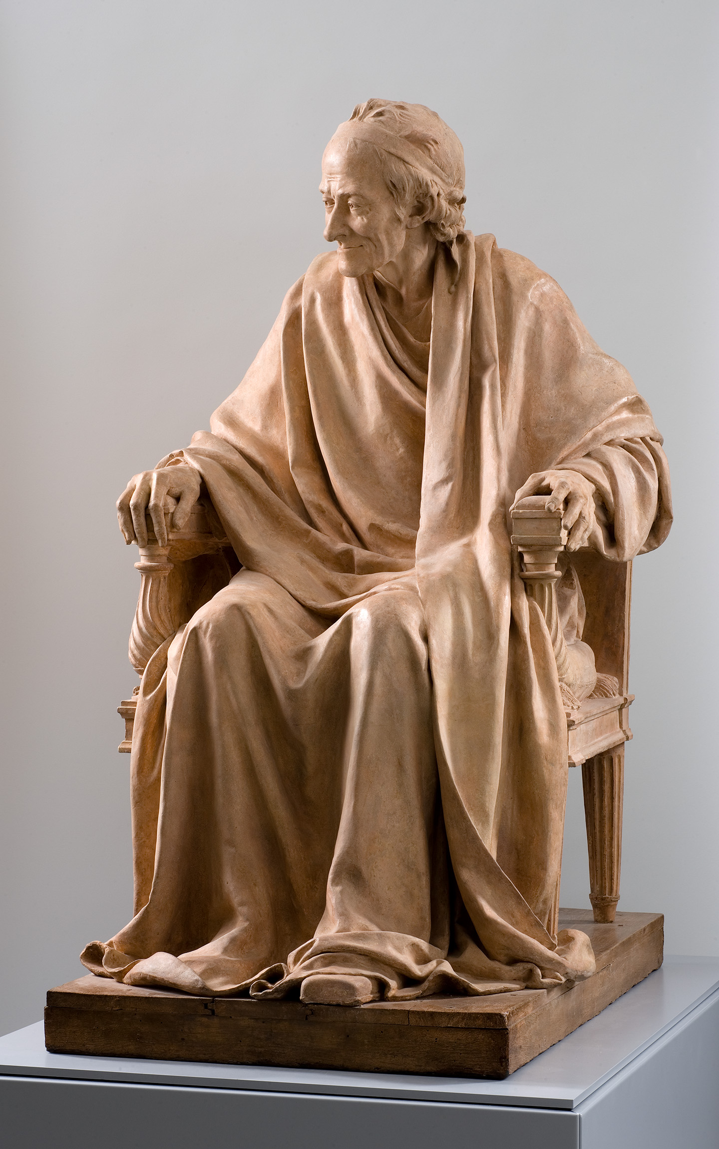 Cette image montre une sculpture en terre cuite représentant un homme assis, vêtu d'une robe ample. Il est placé sur une chaise avec un dossier, ses mains reposant sur les bras de la chaise. Le visage de la sculpture est expressif, affichant une certaine sagesse et sérénité. Les détails de la robe et de la chevelure sont soigneusement réalisés, donnant une impression de réalisme. L'ensemble de la sculpture évoque une attitude réfléchie et contemplative.