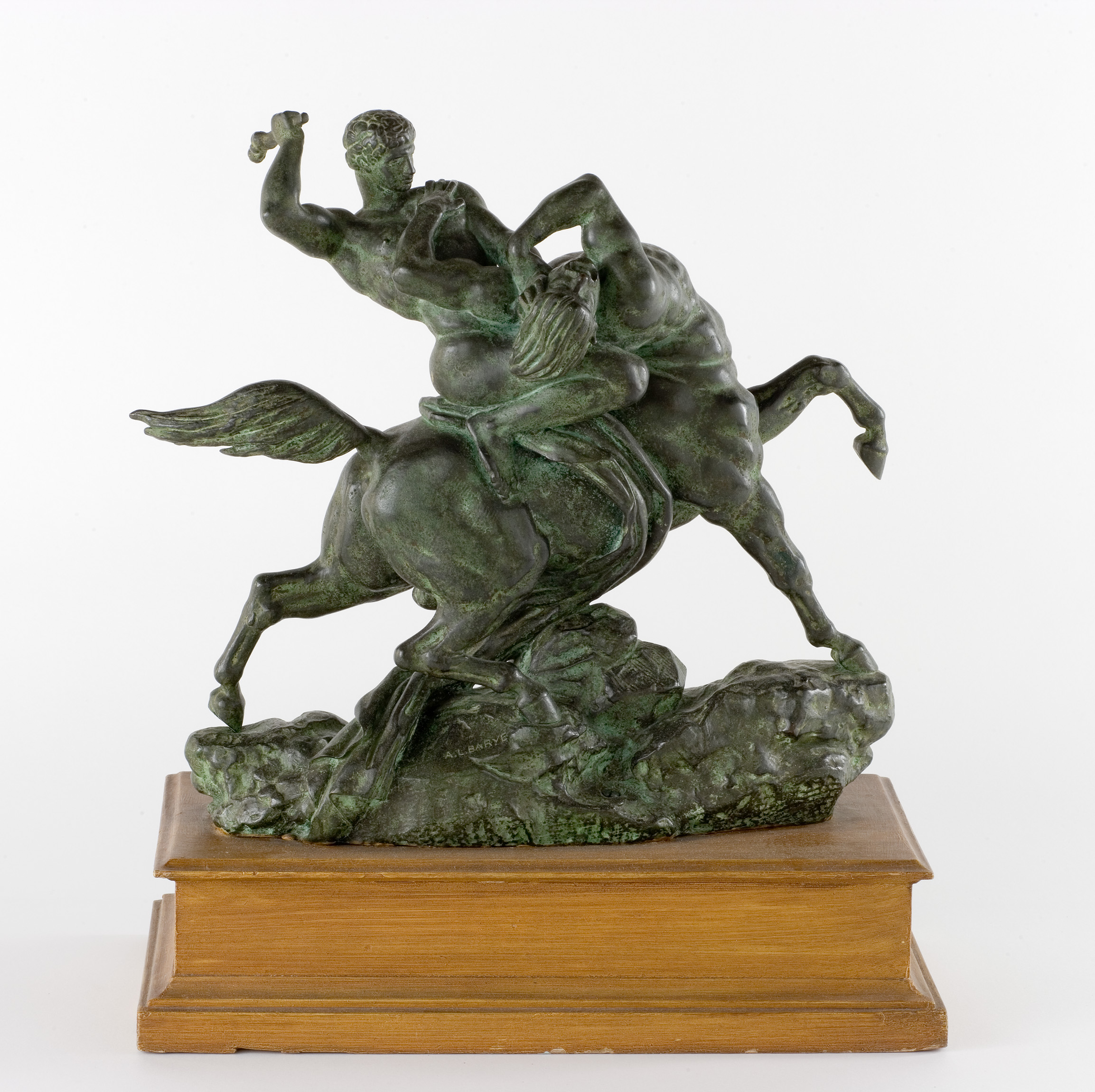 L'image représente une sculpture en bronze de deux figures, un homme et un cheval, en pleine lutte. L'homme semble s'agripper à la jambe du cheval, tandis que celui-ci se retourne, créant une dynamique de mouvement intense. La sculpture est placée sur une base en bois, ce qui met en valeur le matériau et les détails de l'œuvre. L'ensemble dégage une impression de puissance et de tension.