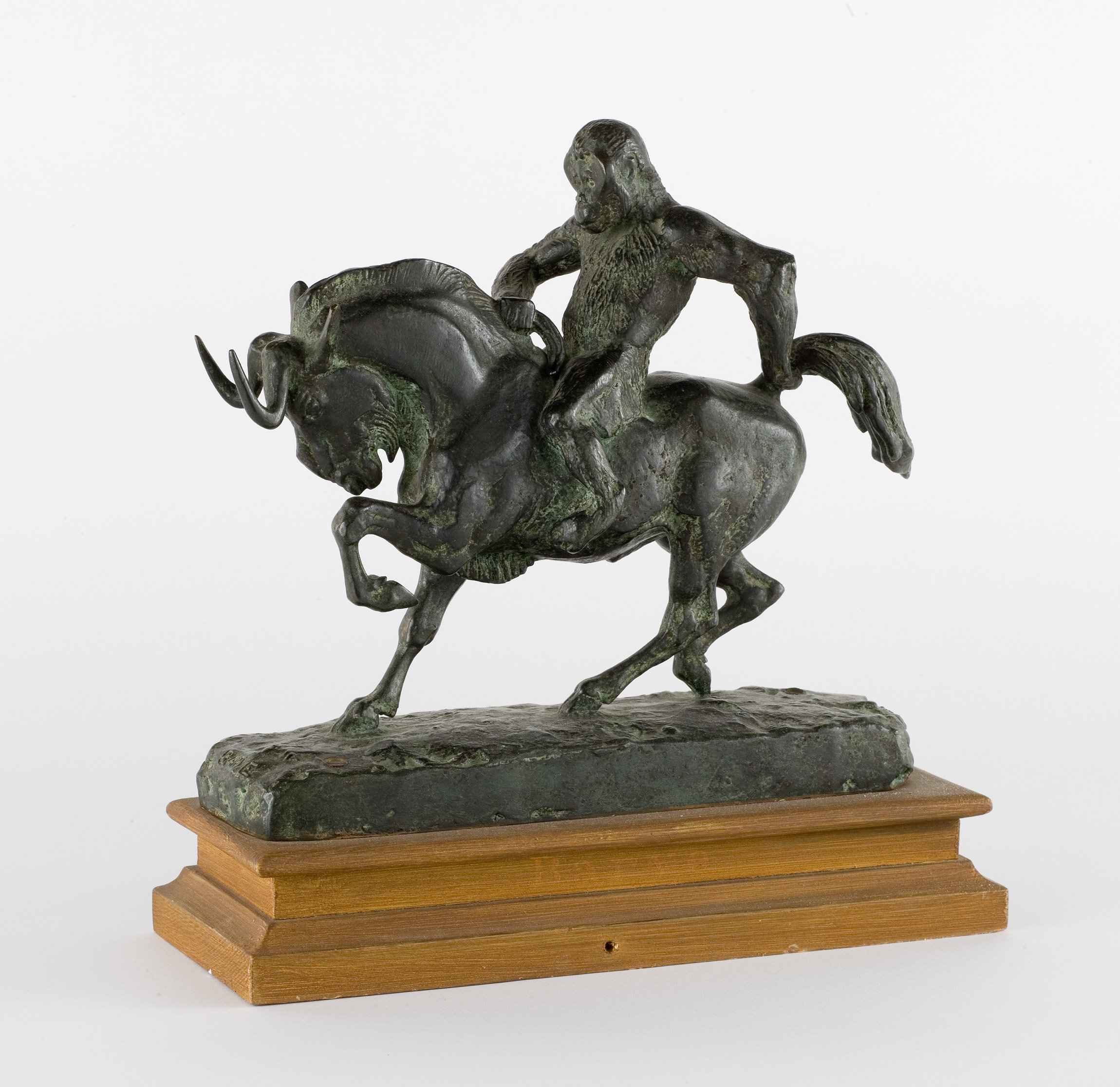 L'image représente une sculpture en bronze d'un cavalier sur un animal, probablement un cheval ou un bétail, en train de galoper. Le cavalier, qui semble avoir une posture dynamique, est représenté avec des détails qui soulignent son mouvement. La sculpture repose sur une base en bois, qui ajoute à son élégance. Ce type d'œuvre pourrait évoquer des thèmes liés à la nature, à l'équitation ou à un contexte historique ou mythologique.