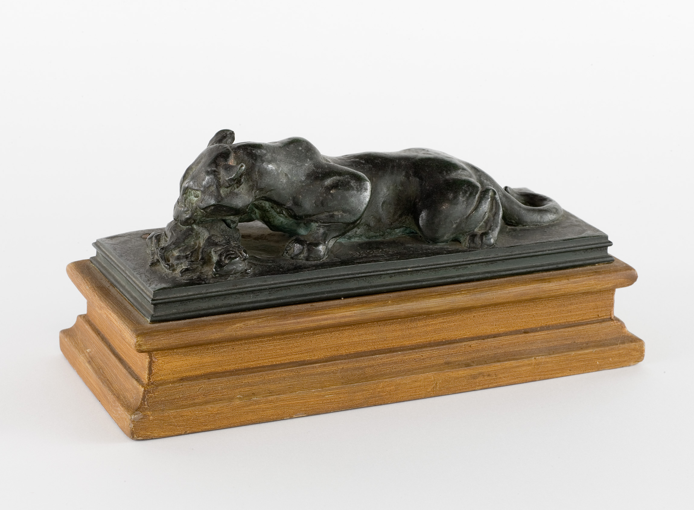 L'image montre une sculpture en bronze représentant un animal couché, probablement un chien, sur une base en bois. Le chien semble être en train de mordre ou de jouer avec un objet, et la sculpture montre des détails réalistes dans sa posture et son pelage. La base en bois est simple, ce qui met en valeur la pièce en bronze. Cette œuvre combine à la fois la finesse du travail du métal et l'élégance du design en bois.