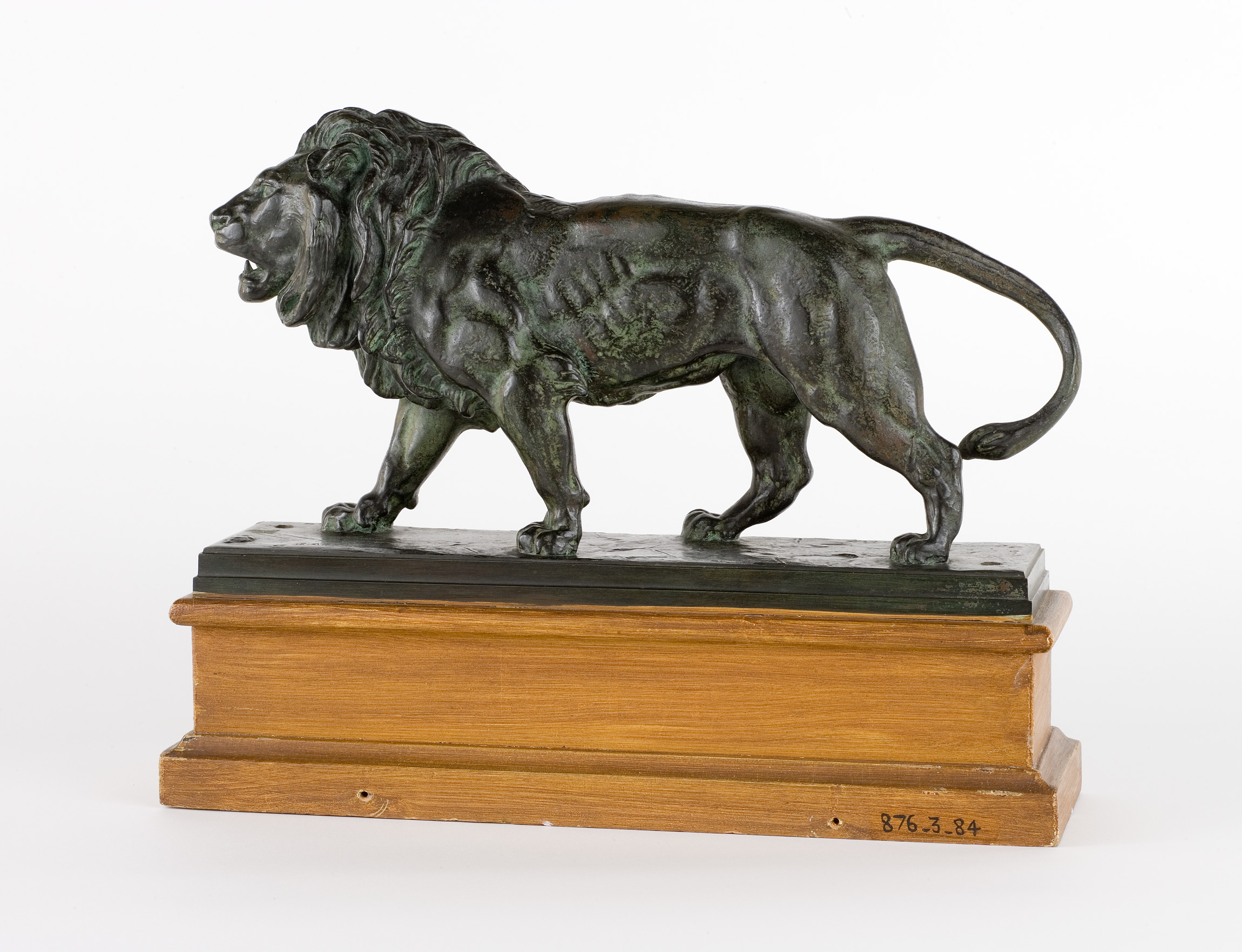 L'image présente une sculpture représentant un lion en bronze. Le lion est debout sur une base en bois, avec une posture fière et majestueuse. Sa crinière est détaillée et ses muscles sont bien définis, ce qui donne une impression de puissance et de mouvement. La sculpture semble être réalisée avec soin, mettant en valeur le réalisme et l'expression animale.