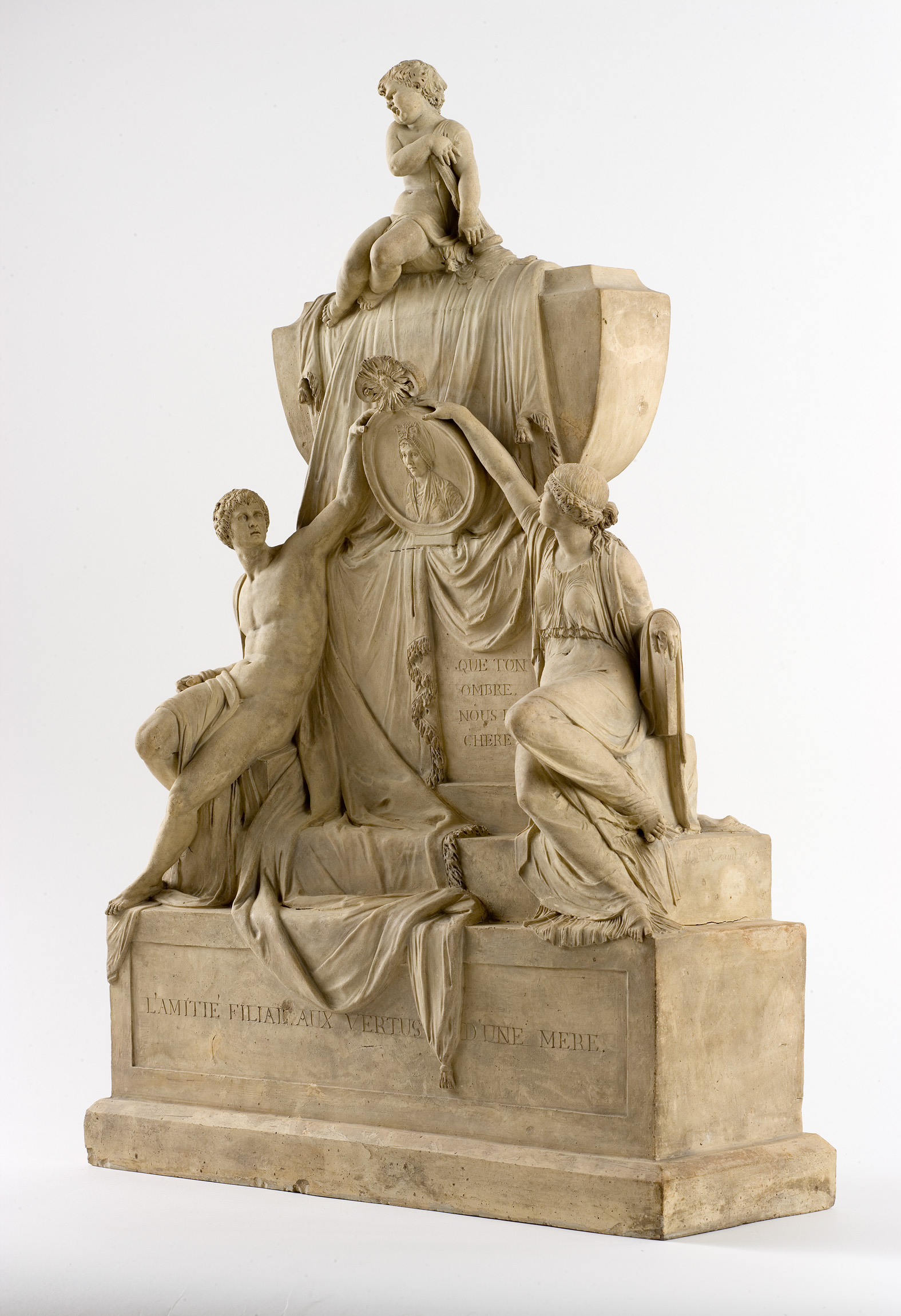 Cette image représente une sculpture en relief de style néoclassique. Au centre, deux figures féminines sont assises, entourées de drapés élégants. L'une d'elles tend une main vers un médaillon qui semble contenir une image, tandis que l'autre est positionnée en arrière-plan, semblant contempler la scène. Une figure enfantine est perchée en haut, ajoutant une touche de légèreté à l'ensemble. La scène évoque des thèmes d'amour filial et de maternité, soulignés par l'inscription "L'amitié filiale envers une mère" qui se trouve sous les personnages. L'œuvre transmet une émotion profonde de tendresse et d'attachement familial.