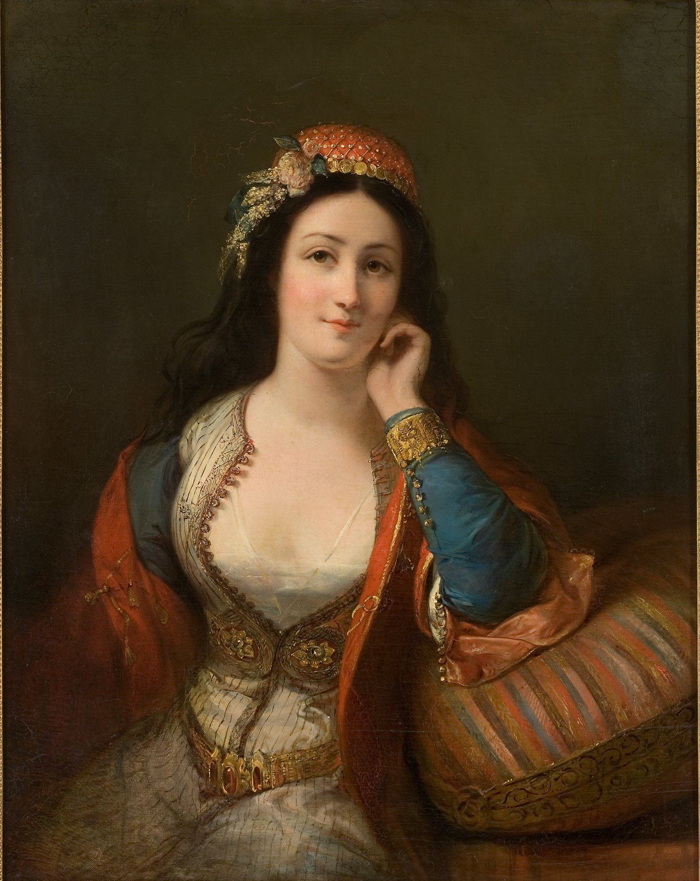 Cette image représente un portrait d'une femme élégante, probablement d'origine méditerranéenne. Elle est vêtue d'une tenue ornée, avec des détails de broderies et des couleurs riches. Les cheveux sombres, légèrement ondulés, tombent sur ses épaules, et elle porte un couvre-chef décoratif. Son expression est douce et sereine, et elle semble contemplative, appuyée sur sa main. En arrière-plan, les couleurs sont sombres et neutres, mettant en valeur les détails de sa tenue et son visage. Un coussin aux motifs colorés est visible à côté d'elle, ajoutant une touche de sophistication à la scène.