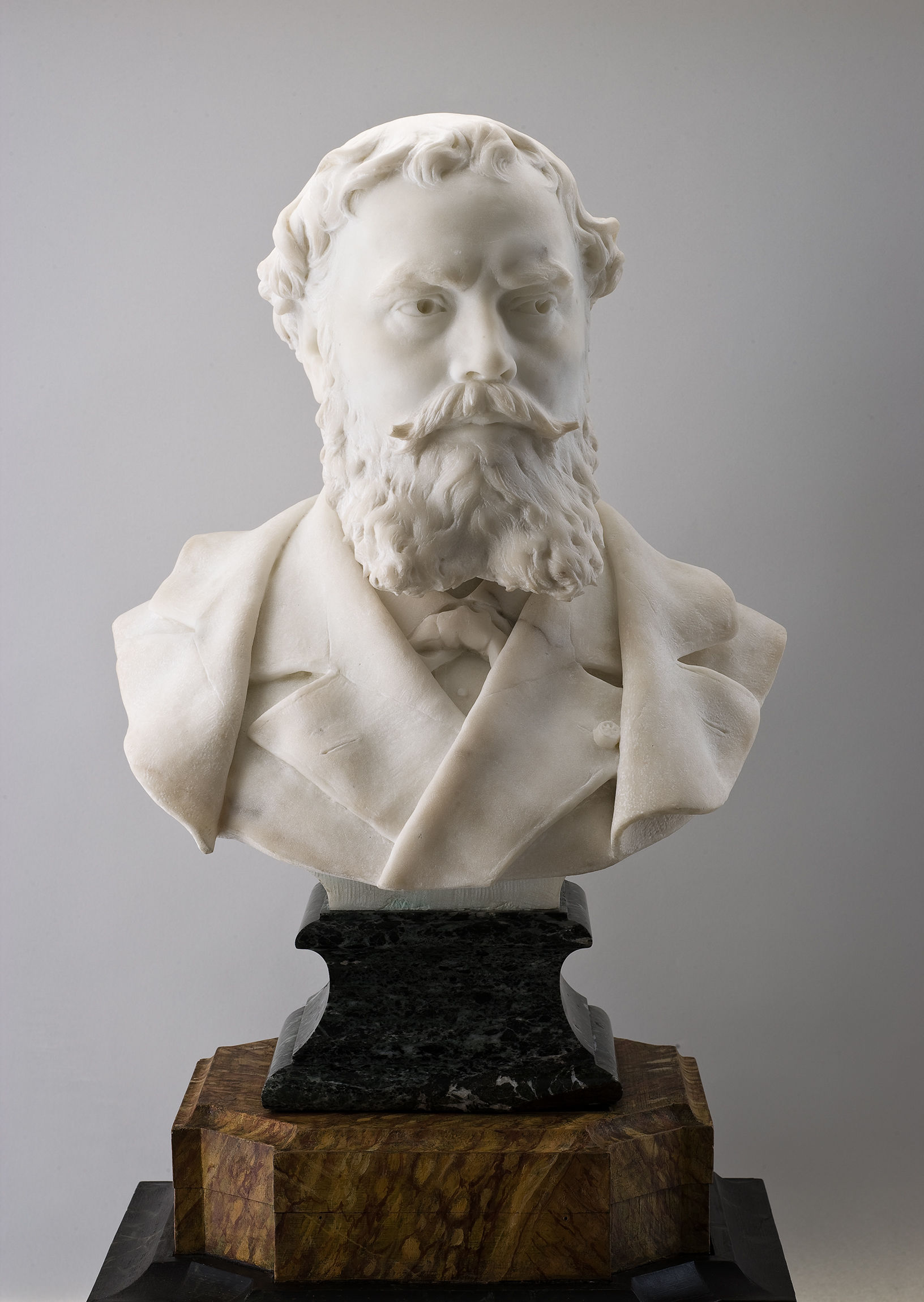 Cette image présente un buste en marbre d'un homme au visage barbu. Le buste est sculpté dans un style réaliste, mettant en valeur les détails du visage et de la barbe. L'homme porte un costume avec un col et une cravate, et le buste repose sur une base en marbre noir et une autre en bois, ajoutant une touche d'élégance à la pièce. L'éclairage met en avant les contours et les textures du marbre, soulignant la maîtrise technique de l'artiste.