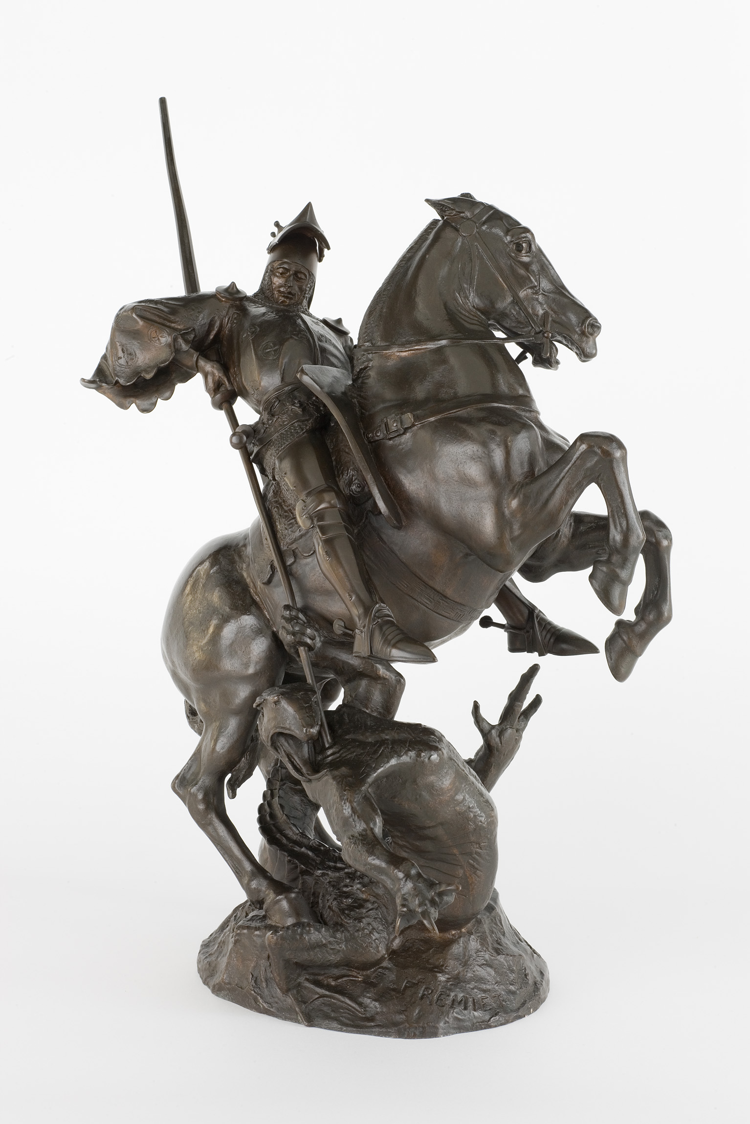 L'image représente une sculpture en bronze d'un cavalier en armure sur un cheval. Le cheval, très dynamique, semble se dresser sur ses pattes arrière, tandis que le cavalier, armé d'une lance ou d'un autre type d'arme, adopte une posture héroïque. La sculpture est riche en détails, montrant les textures de l'armure et le mouvement intense du cheval. À ses pieds, on peut apercevoir une silhouette suggérant une lutte ou un affrontement, ce qui ajoute une dimension dramatique à l'œuvre.