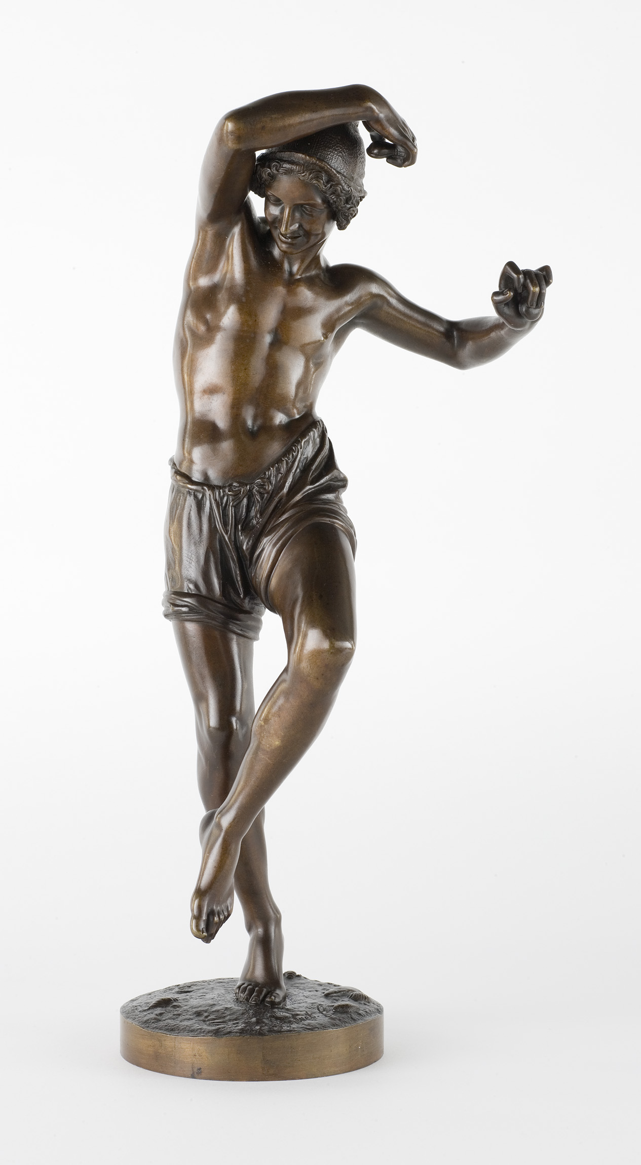 L'image représente une sculpture en bronze d'un jeune homme en mouvement. Il a un torse nu, portant seulement un short. Sa posture est dynamique, avec un bras levé et une jambe légèrement fléchie, comme s'il dansait ou se préparait à sauter. L'expression de son visage montre une certaine joie ou concentration, ajoutant à l'énergie de la scène. La finition du bronze donne une belle texture et un éclat au corps sculpté.