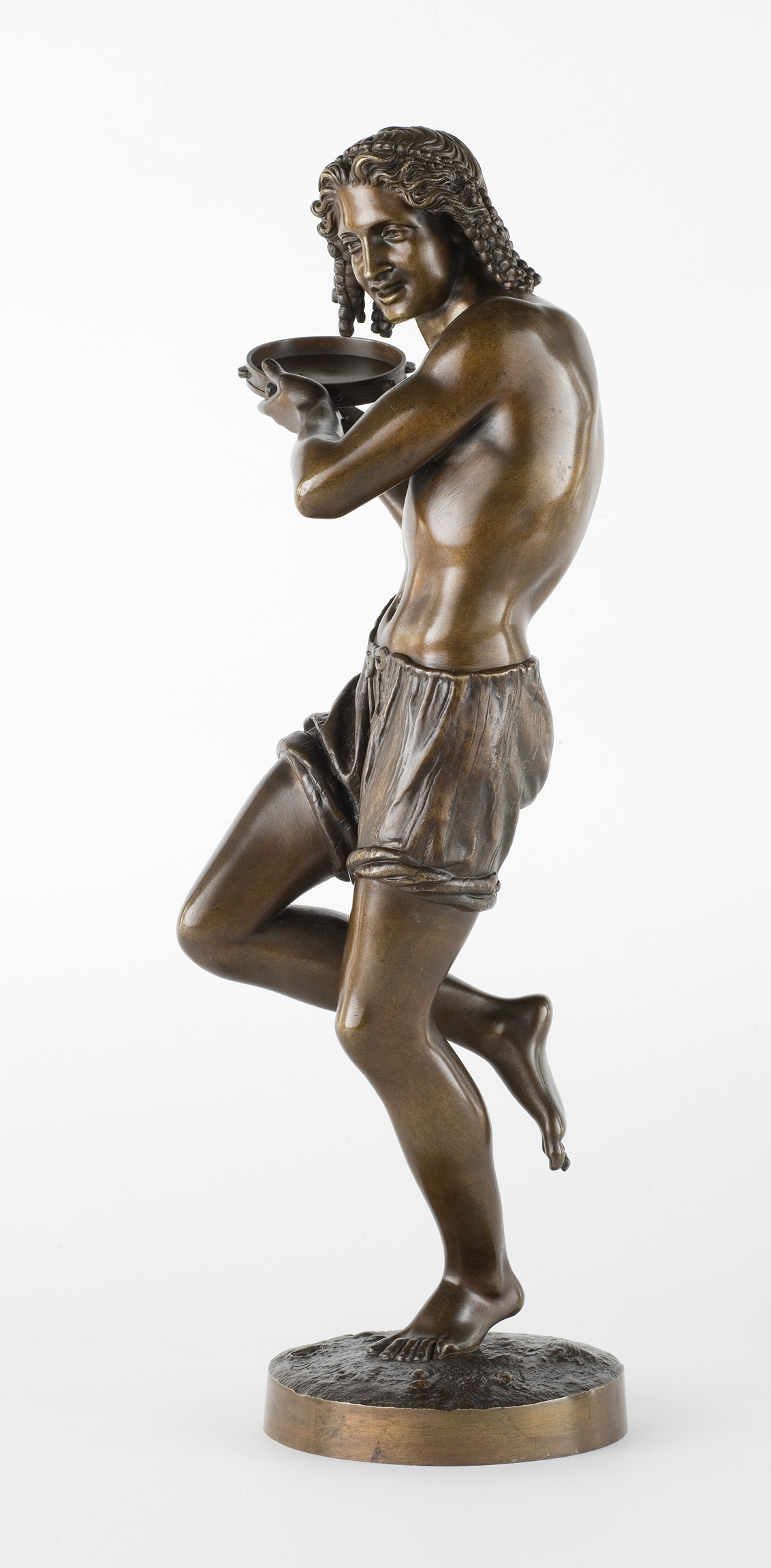 L'image montre une sculpture en bronze représentant un jeune homme nu aux cheveux bouclés. Il se tient en position dynamique, légèrement penché en avant, avec une jambe levée. Ses bras sont repliés, tenant un plat ou un bol. Le détail de la sculpture souligne sa musculature et ses traits, mettant en valeur les textures du bronze. La base de la sculpture est solide, contribuant à l'équilibre de l'ensemble. Cette œuvre suggère une impression de mouvement et de grâce.