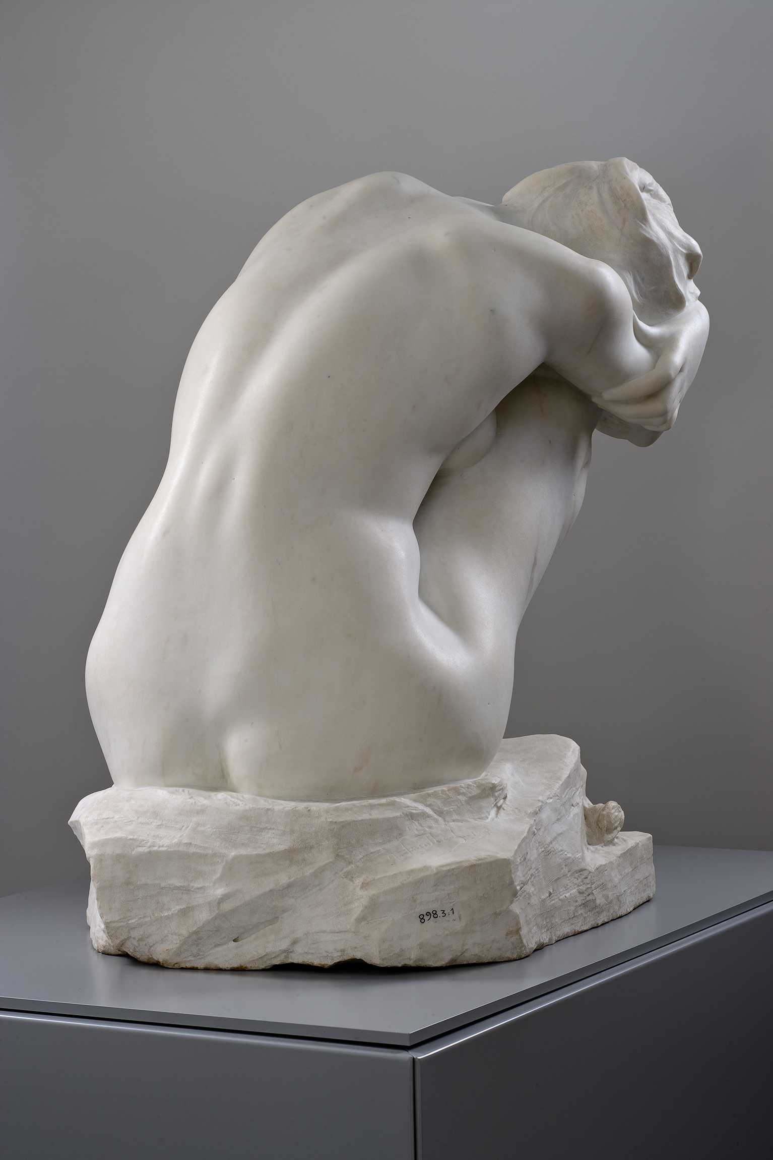 Cette image montre une sculpture en marbre représentant une figure humaine, vue de dos. La sculpture est stylisée et montre une posture de recueillement ou de contemplation, avec le corps légèrement courbé et les bras enroulés autour des genoux. La texture du marbre est lisse, ce qui met en valeur les contours des muscles et les formes du corps. La base sur laquelle repose la sculpture est en pierre, ajoutant à l'aspect solide et intemporel de l'œuvre. Les détails et les ombres suggèrent une grande maîtrise technique de l'artiste.