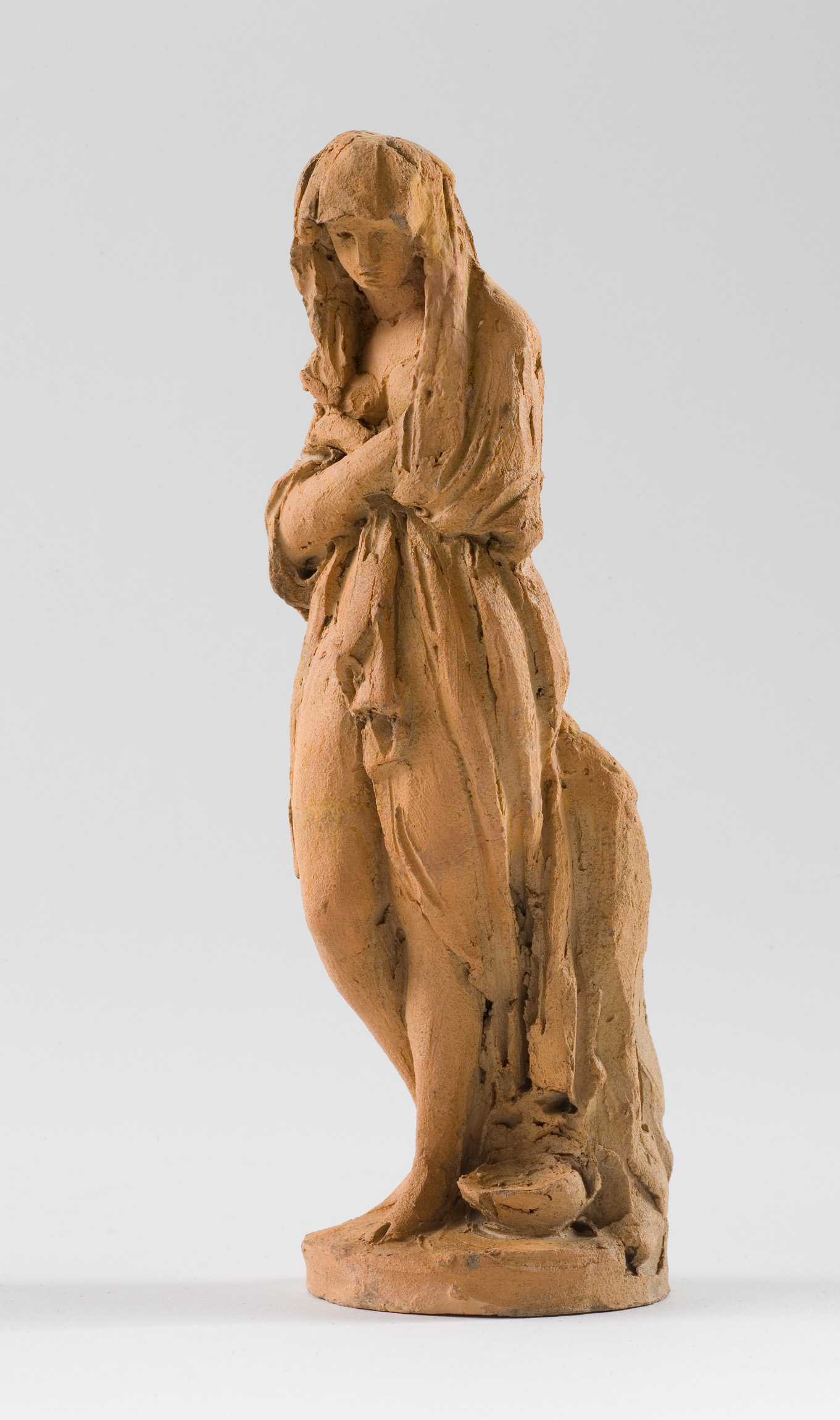 L'image montre une sculpture en terre cuite représentant une femme debout. Elle est drapée dans un vêtement qui semble fluide, avec des plis marqués qui ajoutent du mouvement à la pièce. La femme a les bras croisés et semble pensive ou réservée, avec une expression douce. Ses cheveux sont couverts par un voile, et la sculpture se caractérise par des détails stylisés et une texture rugueuse typique de la terre cuite. L'œuvre évoque à la fois la pudeur et une certaine délicatesse.