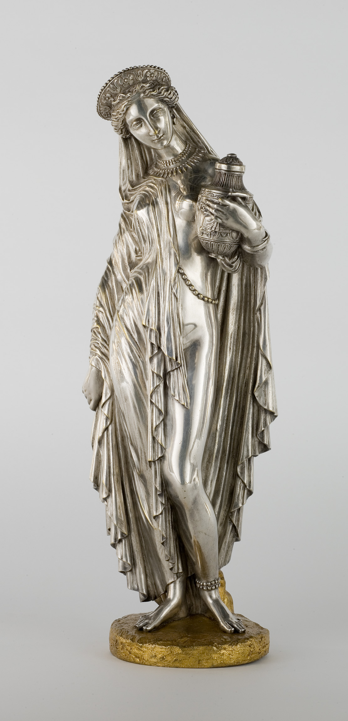 L'image montre une statue représentative d'une figure féminine. La statue est conçue en métal au fini brillant, mettant en valeur des détails raffinés dans sa draperie et ses expressions. La femme porte un drapé élégant qui tombe en ondulations, et elle tient un vase dans une main. Sa posture est gracieuse et elle a une expression douce, évoquant une certaine majesté. La base de la statue est dorée, ajoutant un contraste avec le métal argenté de la sculpture.