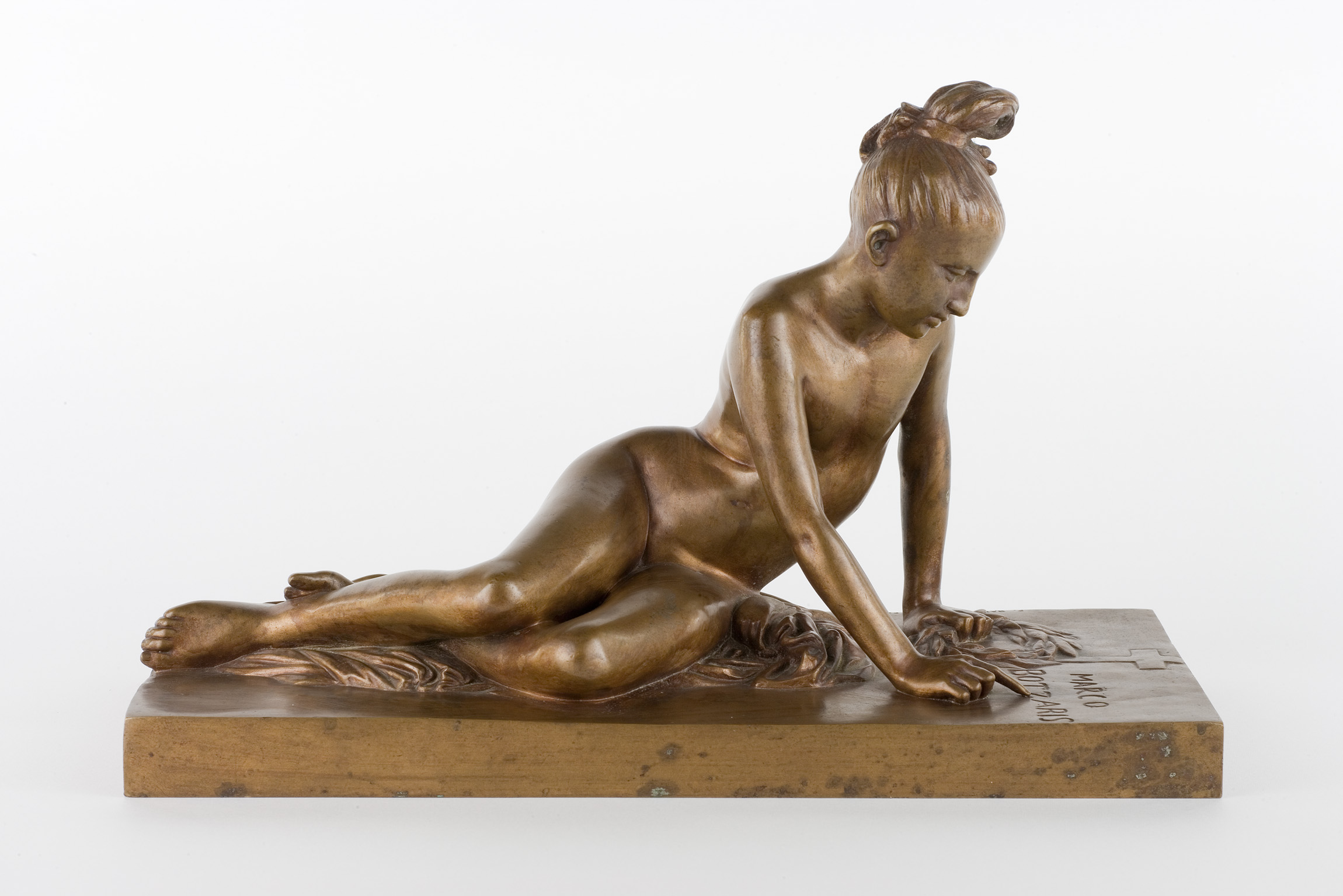 L'image représente une sculpture en bronze d'une jeune femme allongée. Elle est sur le ventre, appuyée sur ses bras, avec une posture décontractée. Sa chevelure est relevée en un chignon, et ses traits sont délicats. Le socle de la sculpture est plat, et on peut y apercevoir une surface texturée, ajoutant à l'esthétique globale de l'œuvre. Les détails du corps sont soigneusement travaillés, mettant en valeur les courbes et la grâce de la figure.