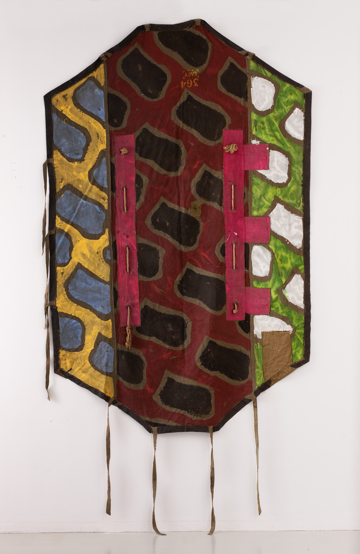 L'image montre un objet artistique de forme octogonale, avec un fond coloré qui présente des motifs géométriques. Les couleurs dominantes incluent des tons de rouge, bleu, vert et jaune, accompagnés de motifs de formes noires. Des accents de rose et de marron sont également présents, ajoutant une dimension texturée à l'œuvre. L'œuvre est probablement un mélange de peinture et de textile, mise en valeur par des attaches en bas qui suggèrent qu'il pourrait s'agir d'une pièce pouvant être accrochée ou portée. Ce type d'œuvre évoque un style inspiré des traditions culturelles, probablement africaines, en raison des motifs et des couleurs vives.