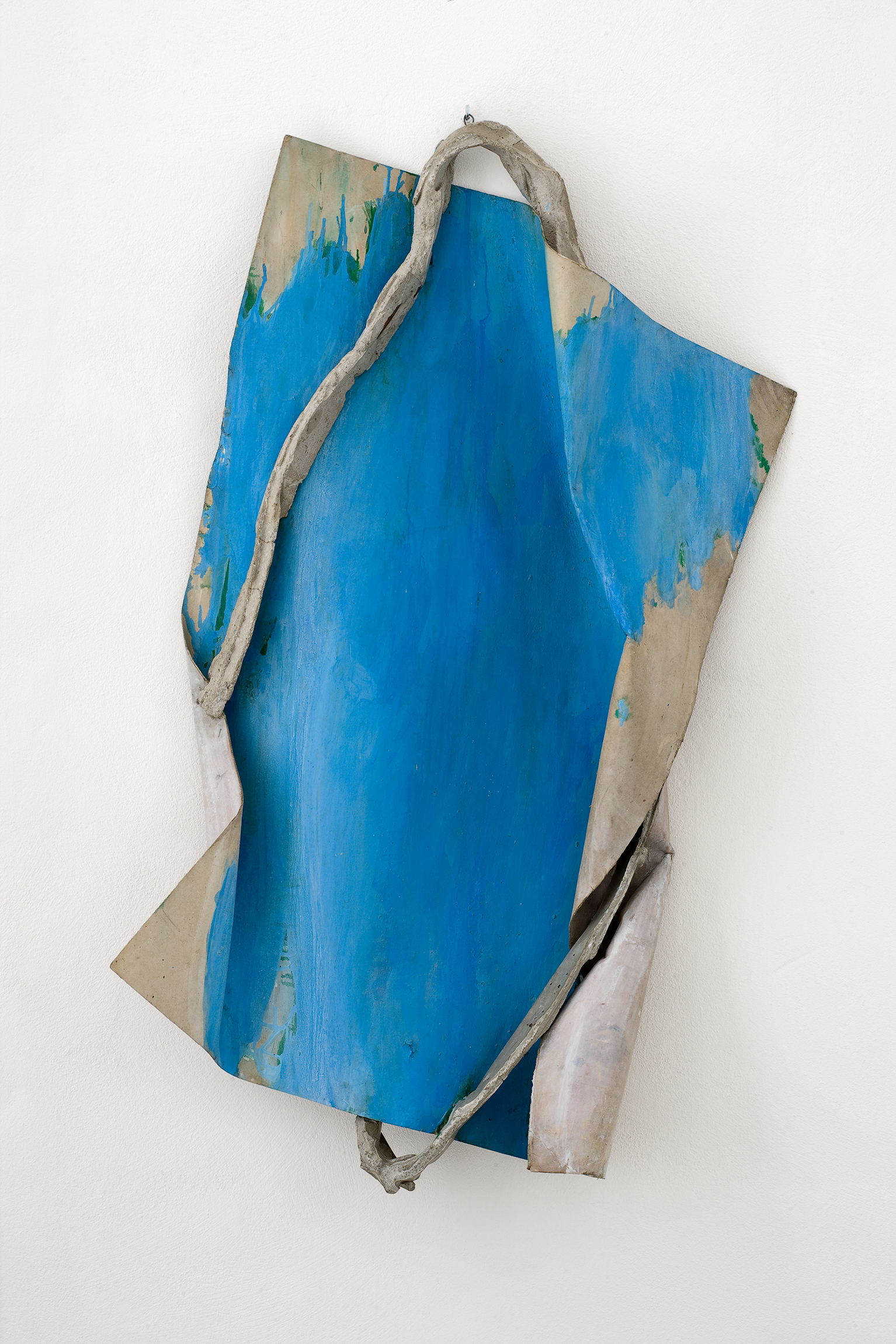 L'image montre une œuvre d'art contemporaine qui se compose d'une forme abstraite aux teintes bleues. La pièce semble être faite de deux matériaux différents : un côté est une surface lisse et brillante de couleur bleue, tandis que l'autre présente une texture plus rugueuse et métallique. Un élément en forme de fil ou de branche entoure partiellement l'œuvre, ajoutant une dimension sculpturale et organique à l'ensemble. L'œuvre est accrochée sur un mur blanc, ce qui met en valeur les couleurs et les textures.