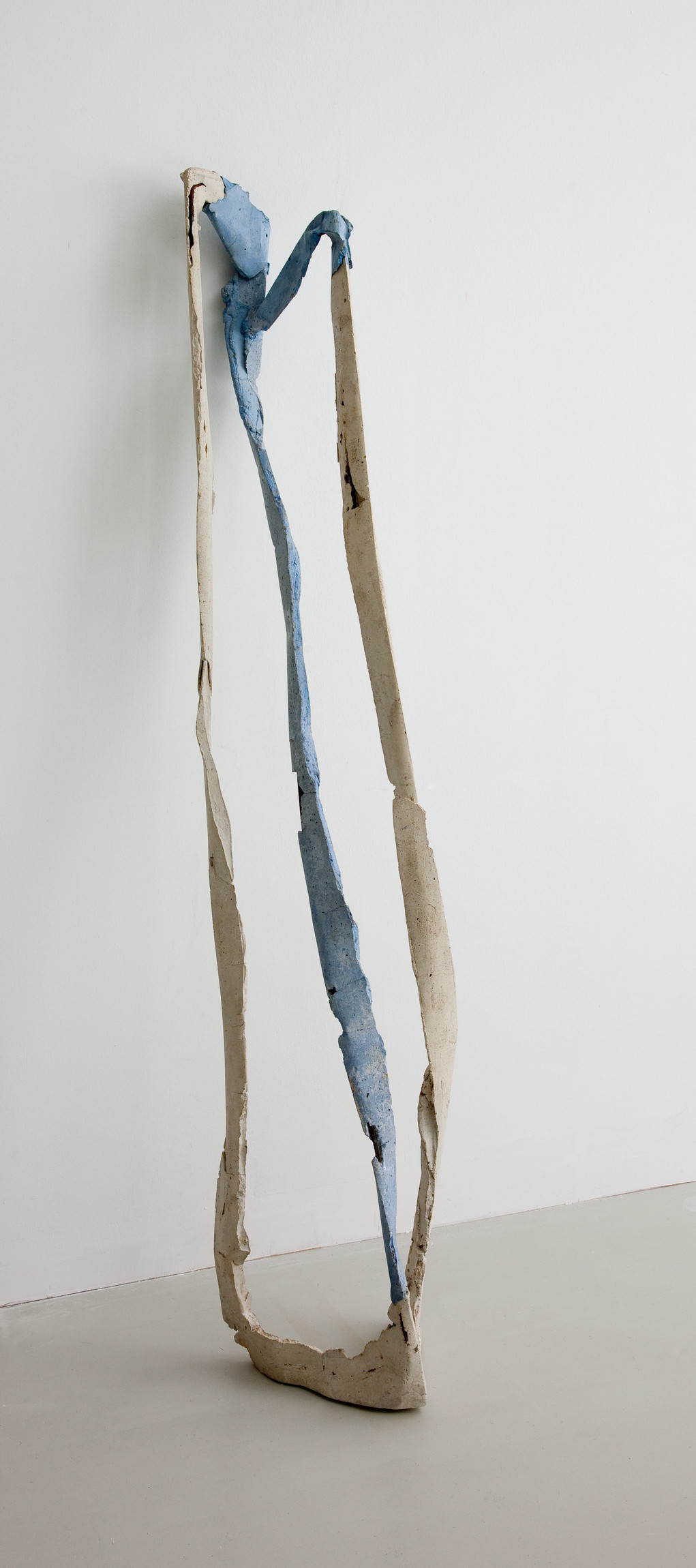 L'image montre une sculpture qui se dresse verticalement. Elle est composée de formes longues et ondulées, avec des sections de couleur beige et bleu. La sculpture semble légère et fluide, avec des plis et des courbes qui lui donnent une apparence organique. Elle est placée contre un mur blanc, sur un sol de couleur claire, ce qui met en valeur la simplicité et l'élégance de l'œuvre. L'ensemble évoque une sensation de mouvement et de légèreté.