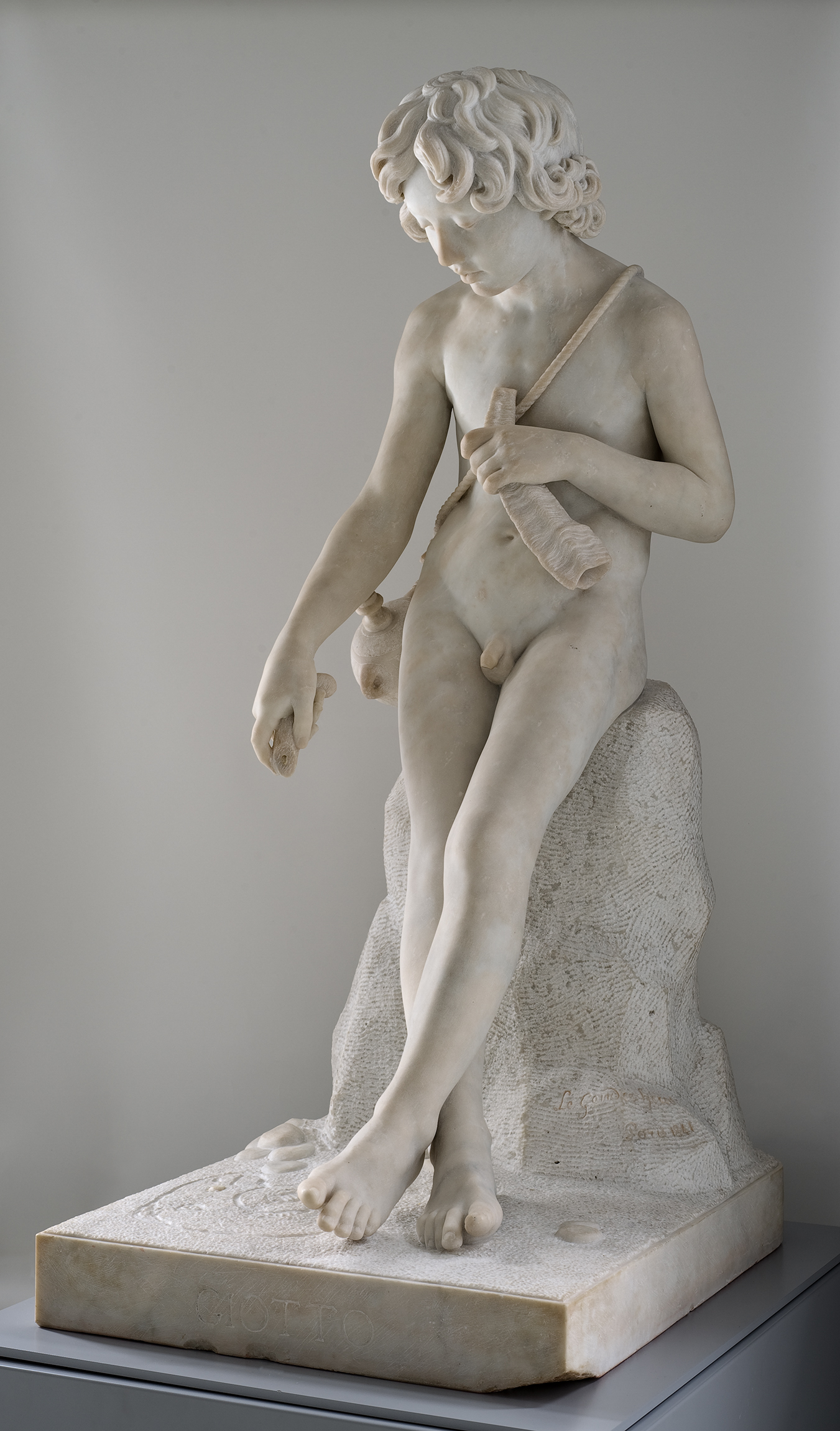 Cette image représente une sculpture en marbre, montrant un jeune garçon nu assis sur un rocher. Il a des cheveux bouclés et il tient un instrument de musique dans une main. Ses pieds sont nus et il semble contemplatif, regardant quelque chose à ses pieds. La sculpture met en avant des détails délicats et un travail minutieux du marbre, soulignant la beauté et la grâce de la figure. L'ensemble évoque une atmosphère tranquille et réfléchie.