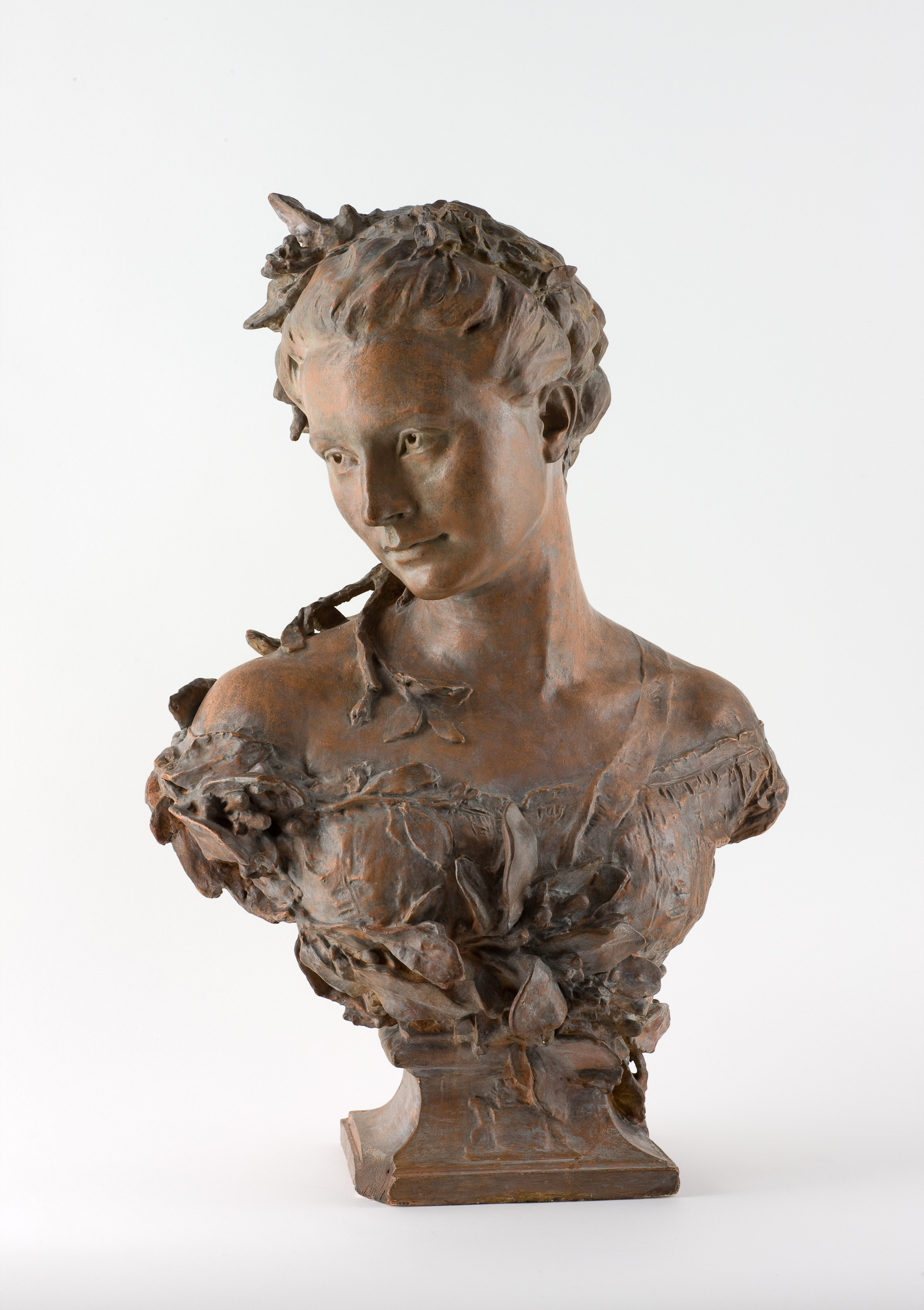 L'image représente une sculpture d'une jeune femme, probablement en bronze, qui montre une attention remarquable aux détails. La statue est partiellement érigée, mettant en avant le buste de la femme. Son visage exprime une douceur et une introspection, avec des traits délicats et un léger sourire. Elle est ornée de feuilles et de fleurs, ajoutant une touche naturelle et romantique à l'œuvre. Le style de la sculpture évoque une époque artistique classique, soulignant à la fois la beauté féminine et l'harmonie avec la nature.