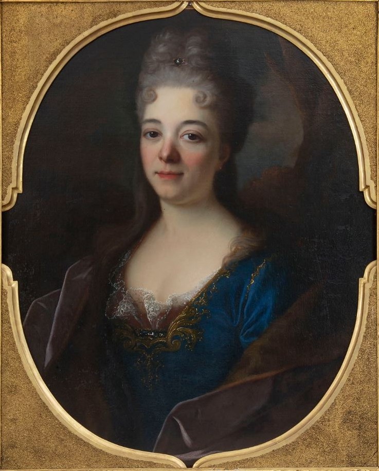 L'image représente un portrait d'une femme du XVIIIe siècle. Elle a des cheveux relevés dans un style élégant, avec quelques boucles et un ornement au niveau du front. Son regard est doux et légèrement détaché, et elle porte une robe bleu richement ornée avec des détails en dentelle. Le fond est sombre, ce qui met en valeur le visage et la tenue de la femme. Le cadre est doré, ajoutant une touche de sophistication au tableau.