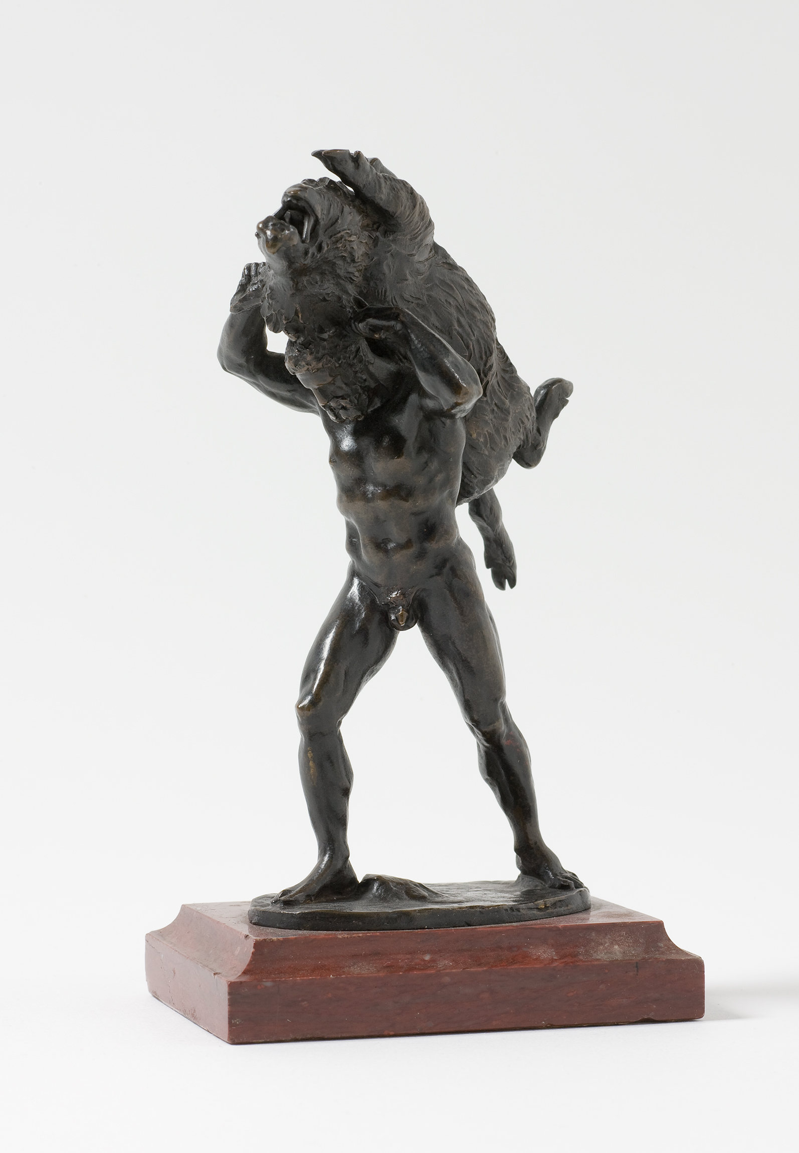 L'image représente une sculpture en bronze d'un homme nu portant sur son épaule un animal (possiblement un mouton ou une chèvre). L'homme a une apparence musclée et semble en position dynamique, comme s'il était en mouvement. La sculpture est fondue sur un socle en bois, ce qui met en valeur le contraste entre le métal et le matériau du socle. Le style de la sculpture évoque une certaine grandeur et une force physique.