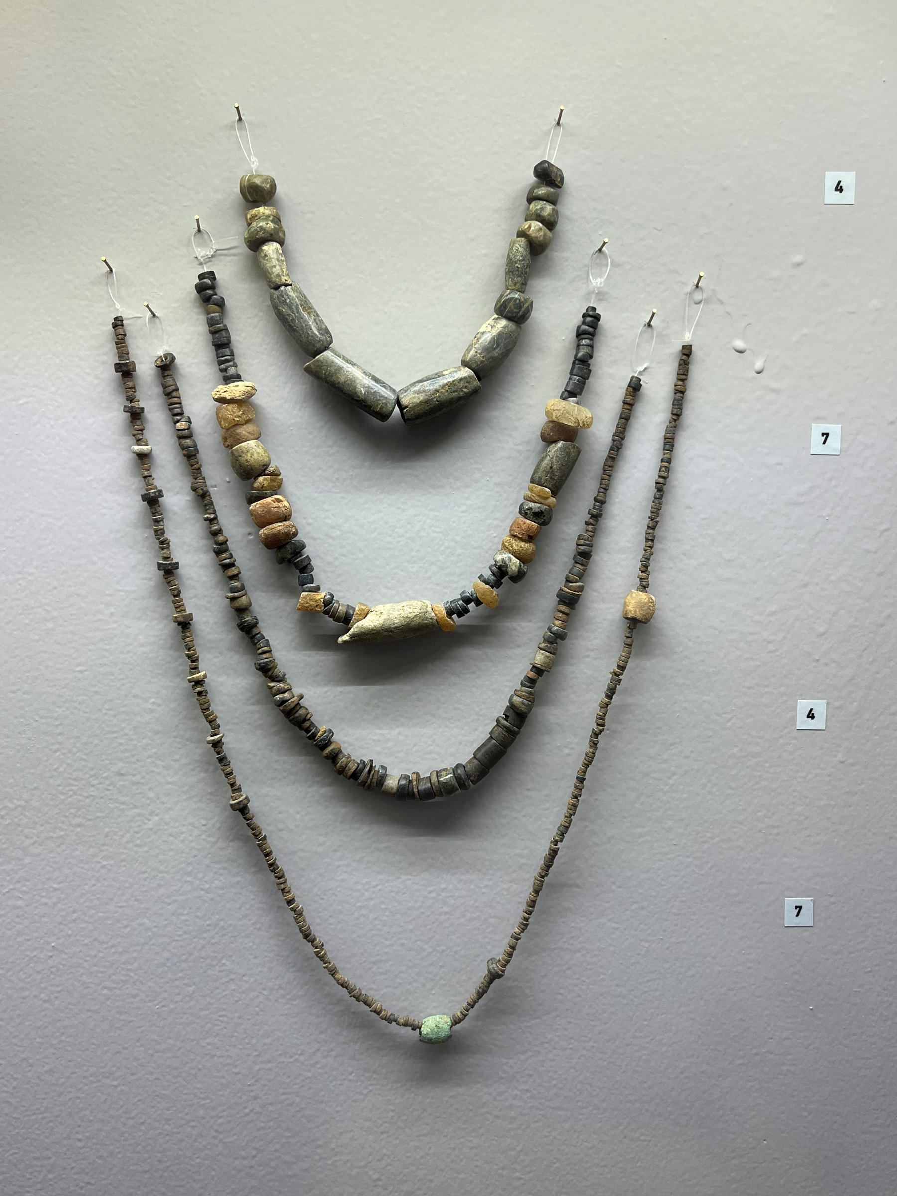 L'image montre plusieurs colliers disposés sur un fond gris. Il y a trois chaînes en haut, composées de perles de formes et de couleurs variées, y compris des perles de pierre et de terre cuite. Les colliers sont de longueurs différentes et paraissent anciens, témoignant d'un savoir-faire artisanal. Ils sont accrochés avec des crochets, ce qui suggère qu'ils sont exposés dans un musée ou une galerie.