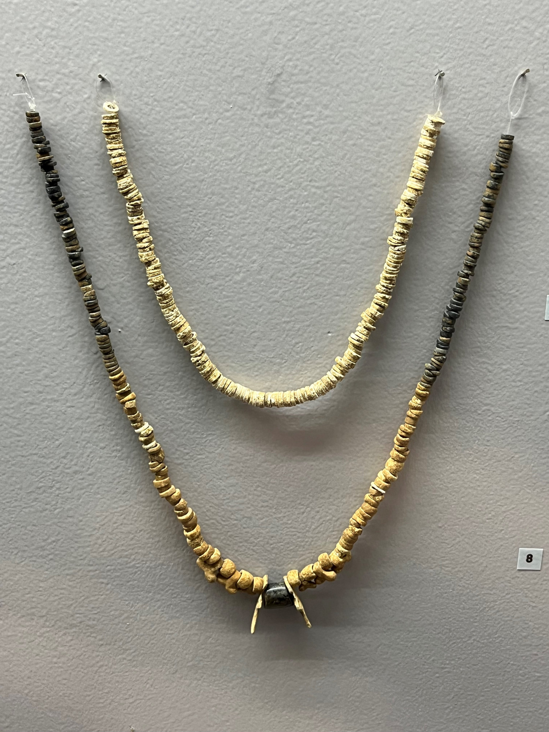 L'image montre deux colliers disposés verticalement sur un mur. Le collier supérieur est constitué de perles de couleur claire, tandis que le collier inférieur présente des perles de couleurs variées, avec des tons allant du beige au noir. Les perles semblent être faites d'un matériau naturel et sont enfilées sur un fil. Ces colliers pourraient avoir une signification culturelle ou artistique, mais cela n'est pas précisé dans l'image.